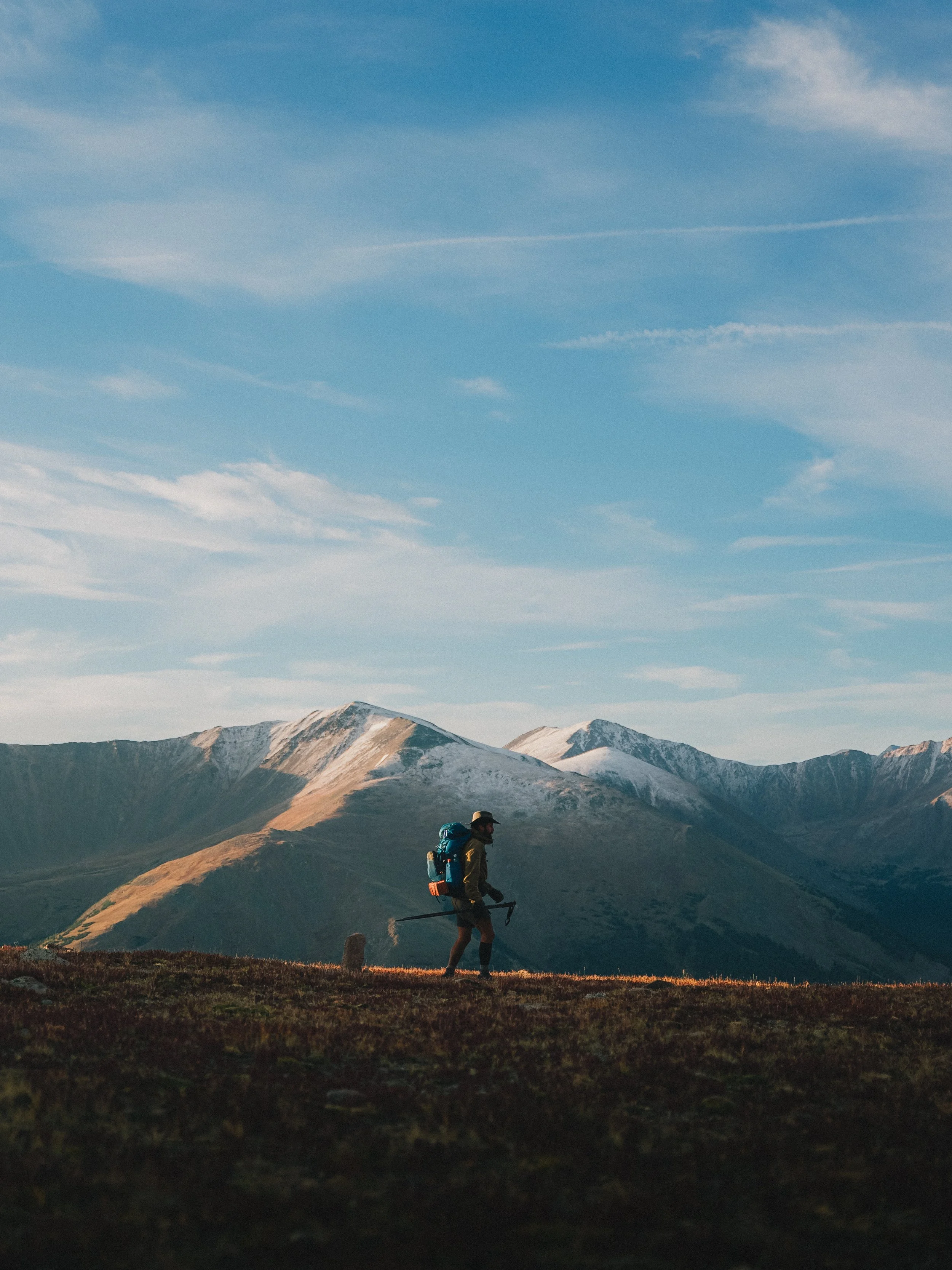 A hiker with a backpack and walking stick trekking through an open grassy area with snow-capped mountains in the background under a blue sky with scattered clouds.