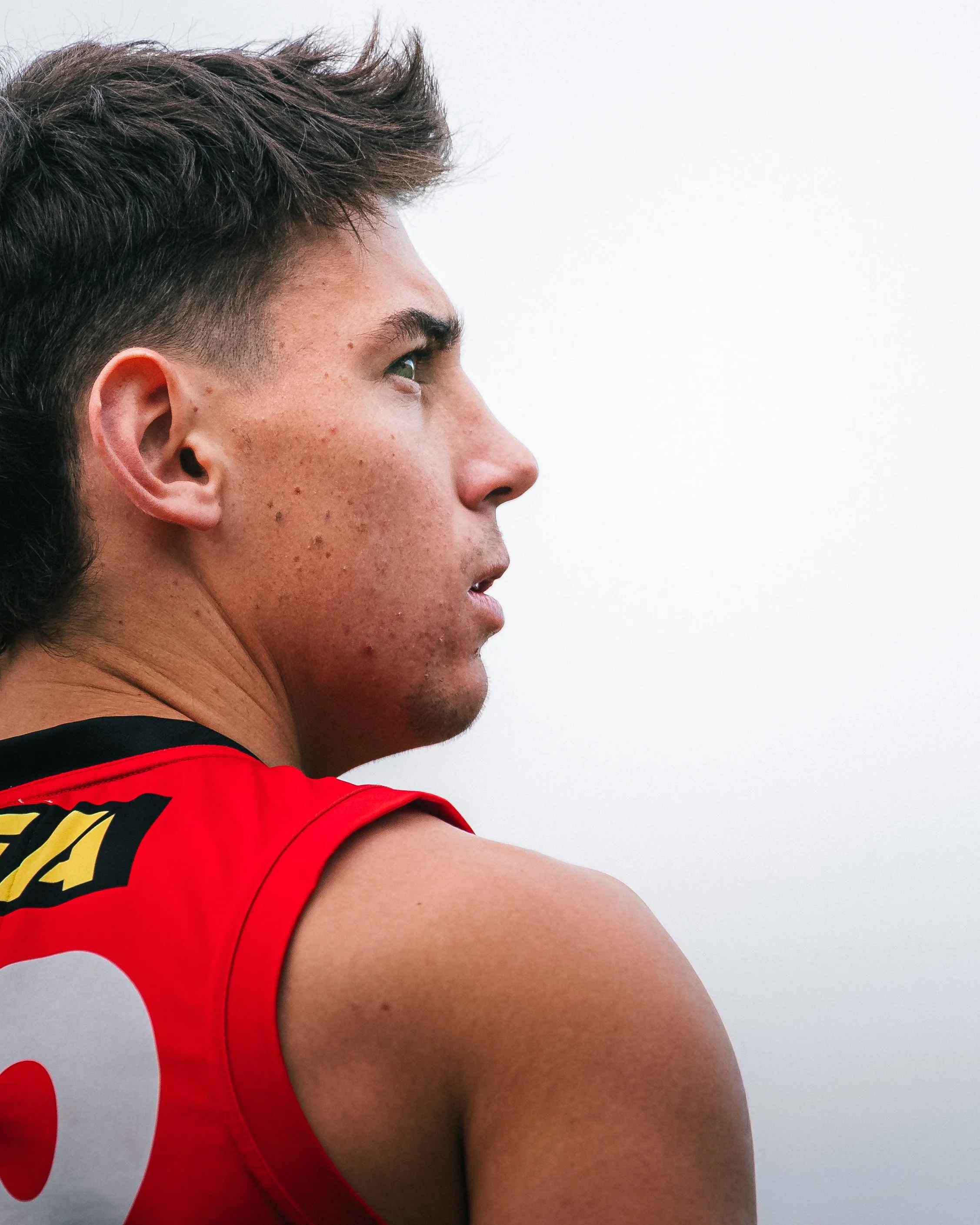Close-up profile of a young man with short brown hair and a tan complexion, wearing a red sports jersey, against a plain light background.