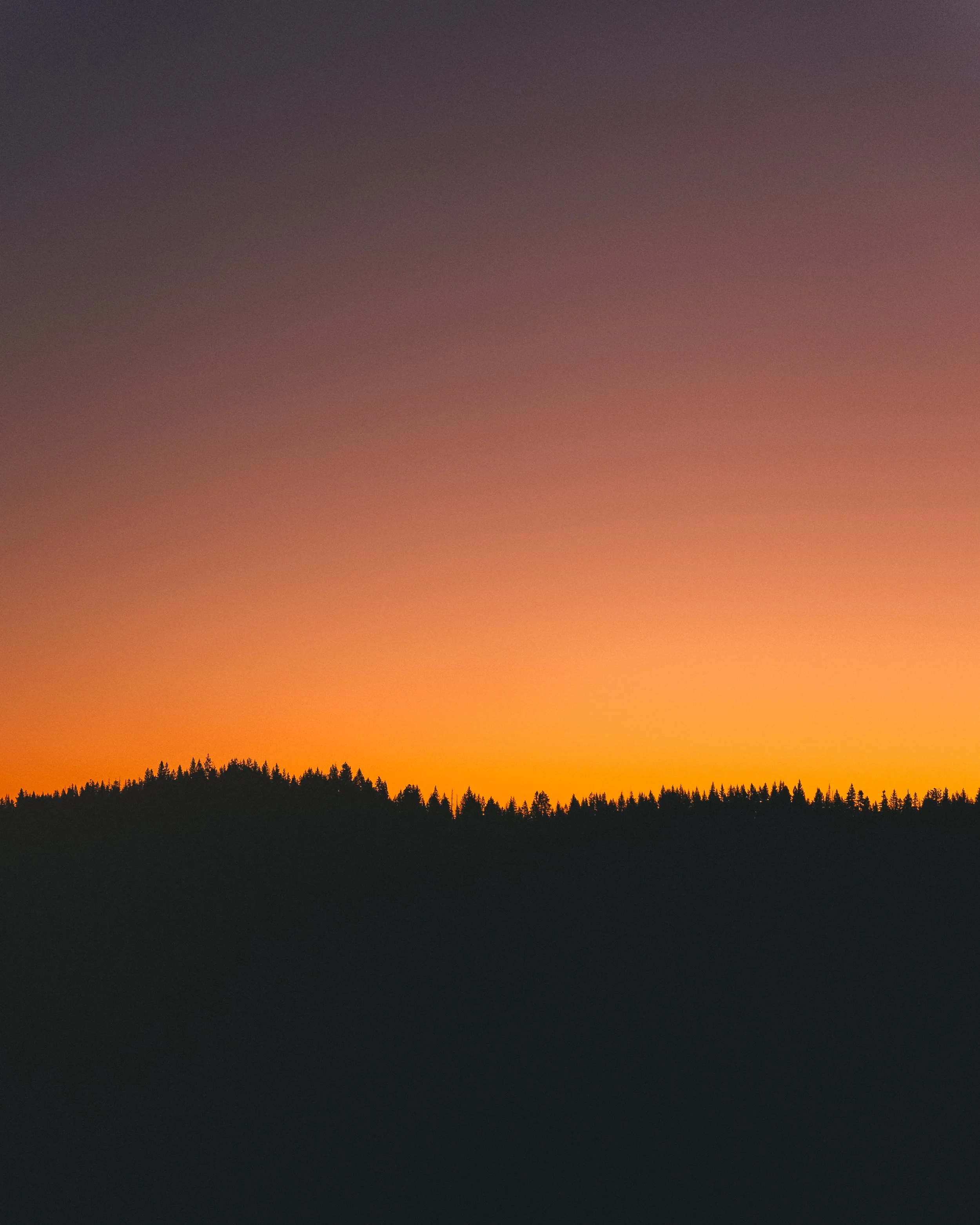 Sunset over a silhouette of a tree-covered hill with an orange and purple sky.
