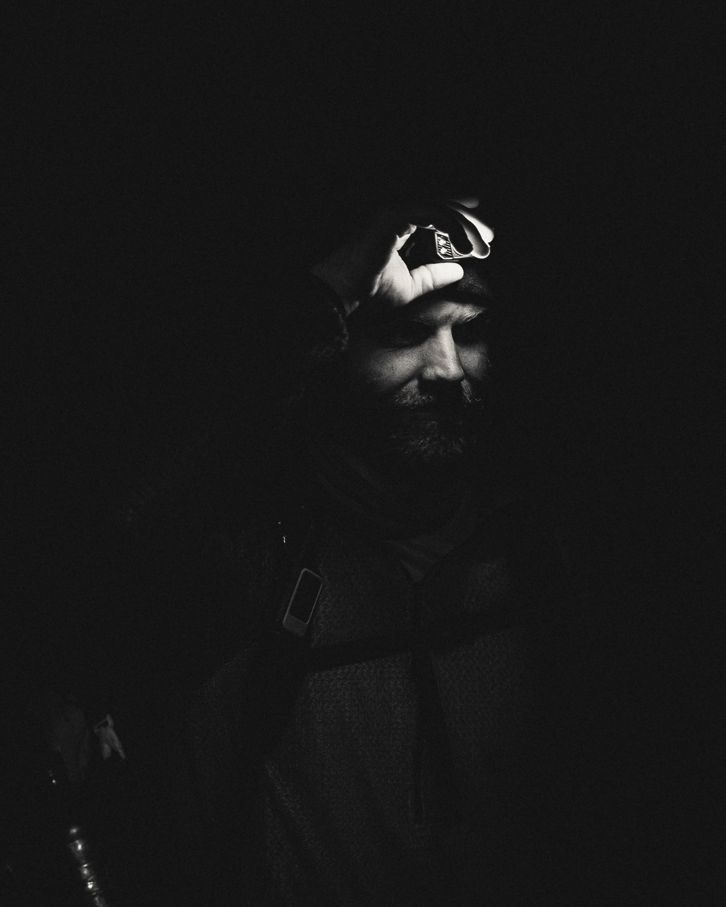 A black and white photo of a man with a beard, holding a phone to his head, with the light illuminating part of his face and hand.