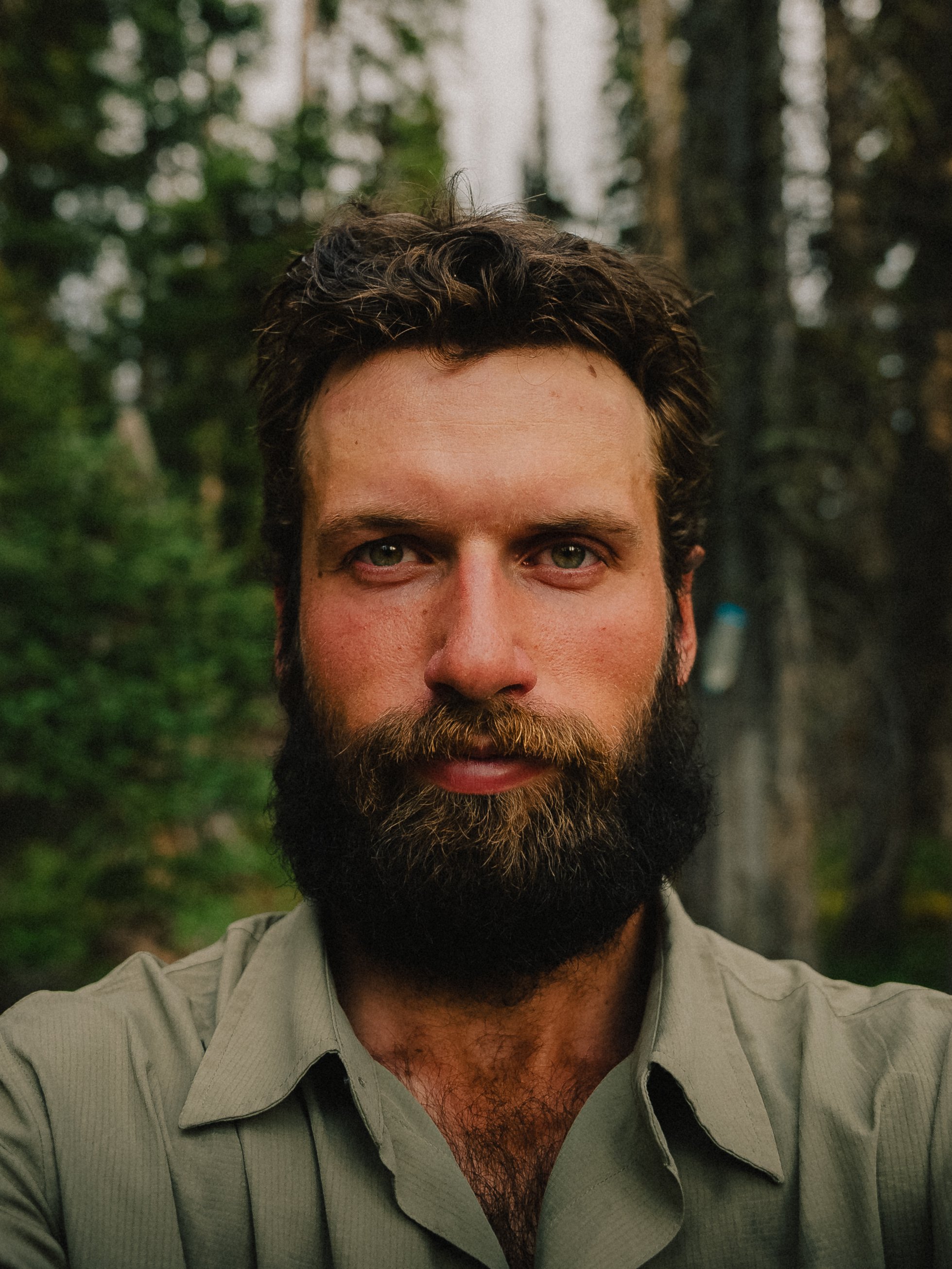 Close-up portrait of a man with a thick beard and green eyes, wearing a light-colored shirt, standing outdoors in a forest with trees in the background.