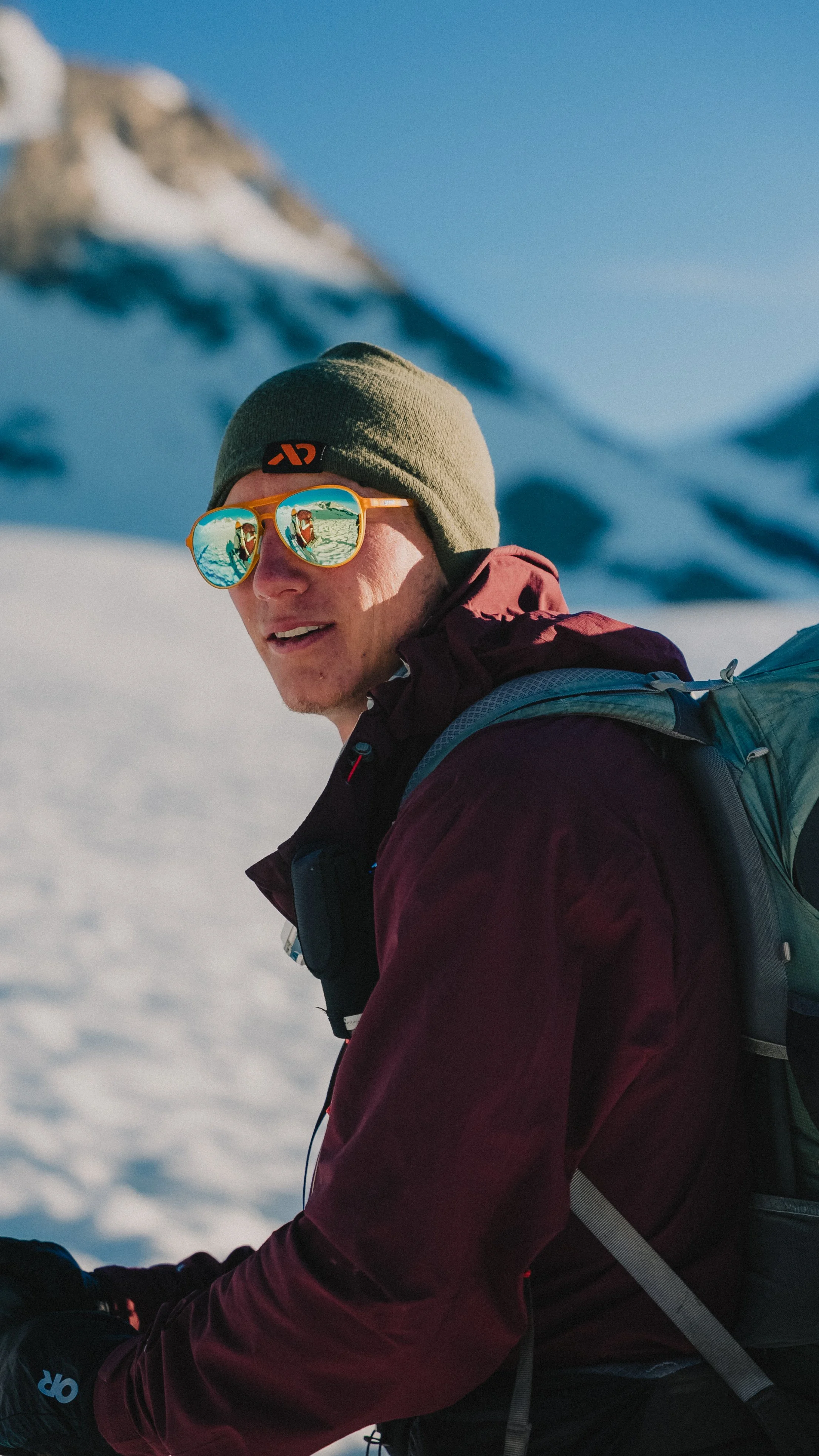 A man in winter gear, sunglasses, and a beanie, standing outdoors in a snow-covered mountainous area.