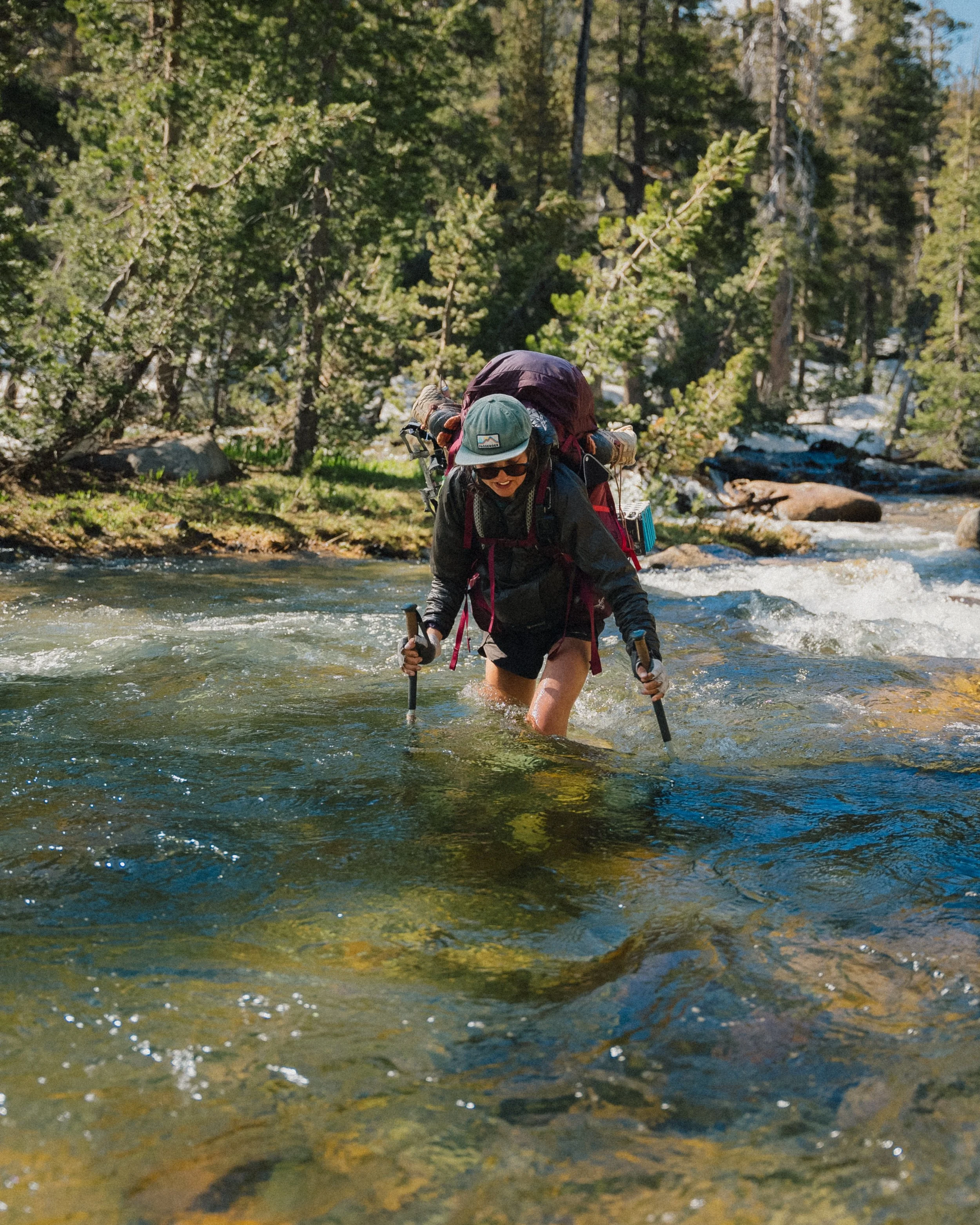 Hiker crossing a rushing river in a forested area, wearing a backpack, sunglasses, cap, and using trekking poles.