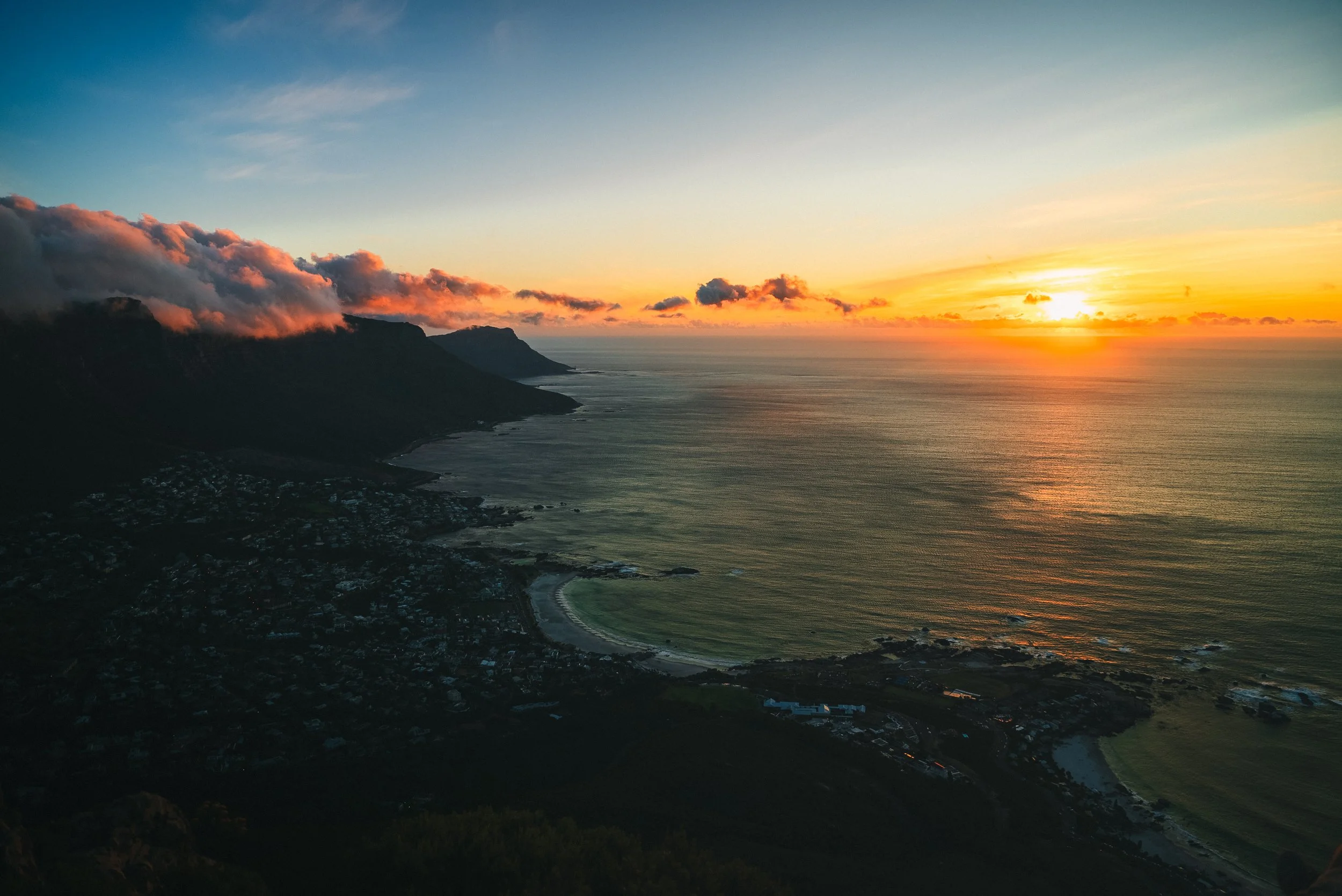 Aerial view of a coastal landscape during sunset, with mountains on the left, a small town near the shoreline, and the ocean extending to the horizon under a partly cloudy sky.
