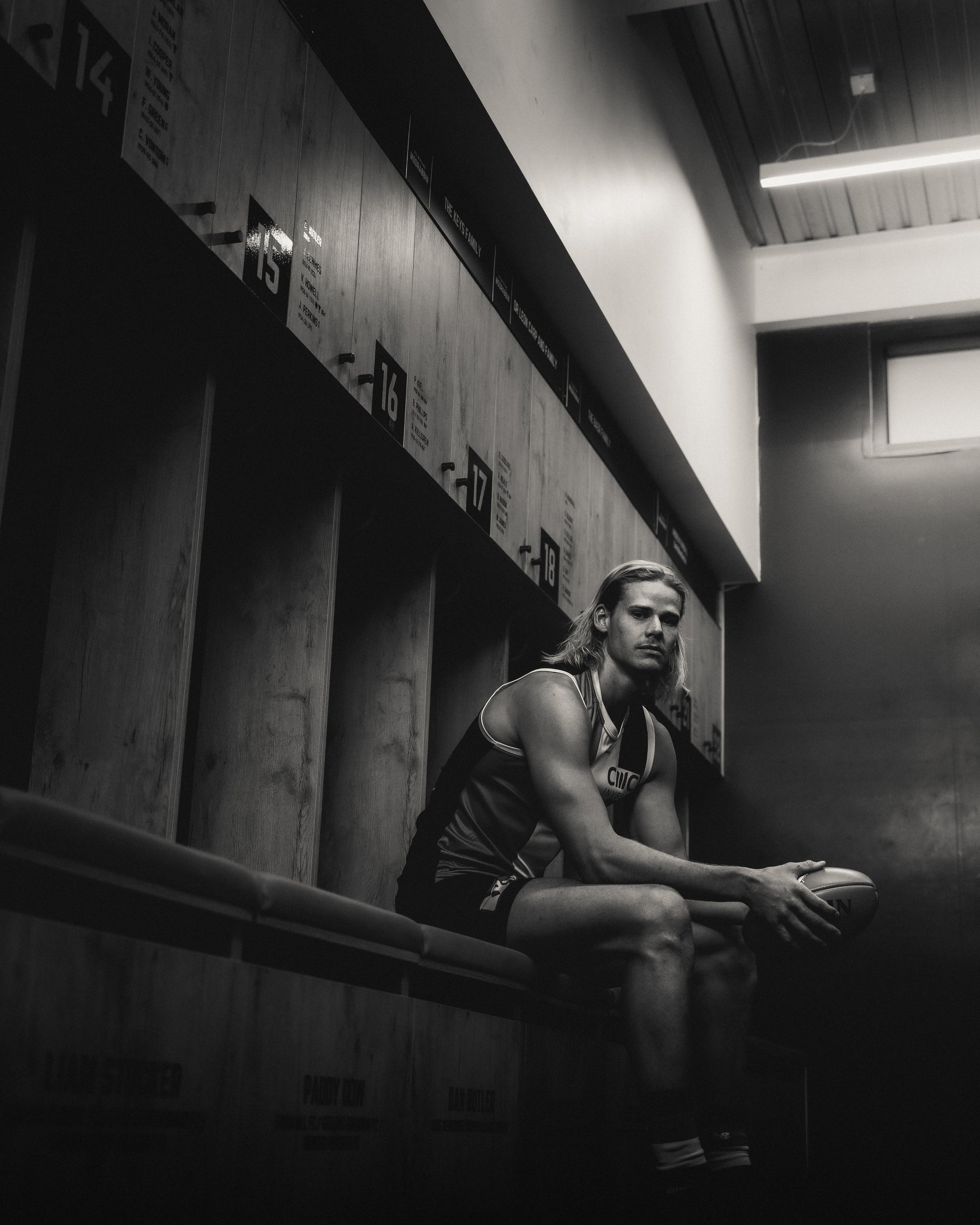 Black and white photo of a male athlete sitting on a bench in a locker room, holding a rugby ball, wearing sports gear, with lockers in the background and a fluorescent light overhead.