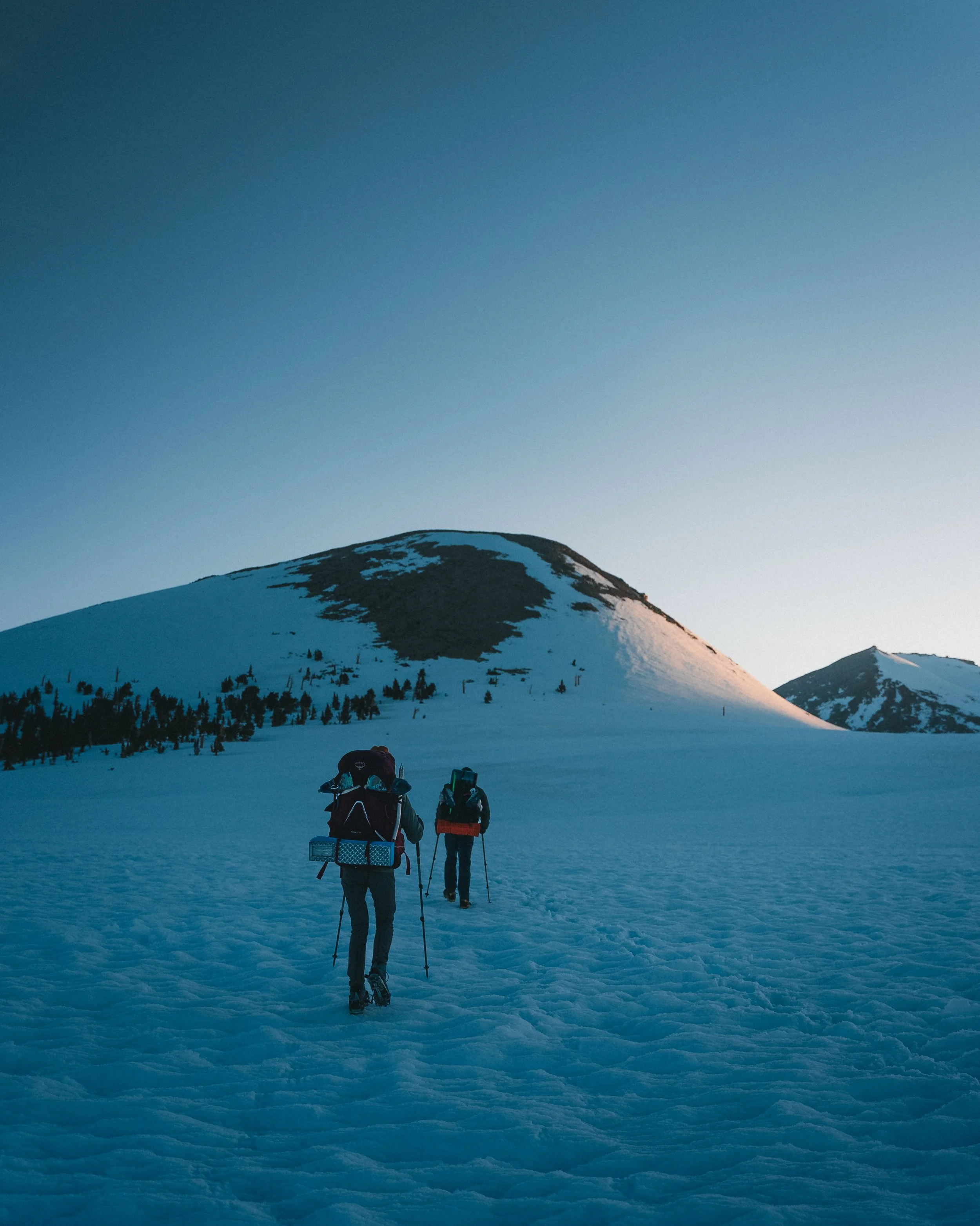 Two hikers with backpacks trekking across a snow-covered landscape towards mountainous terrain during sunset.
