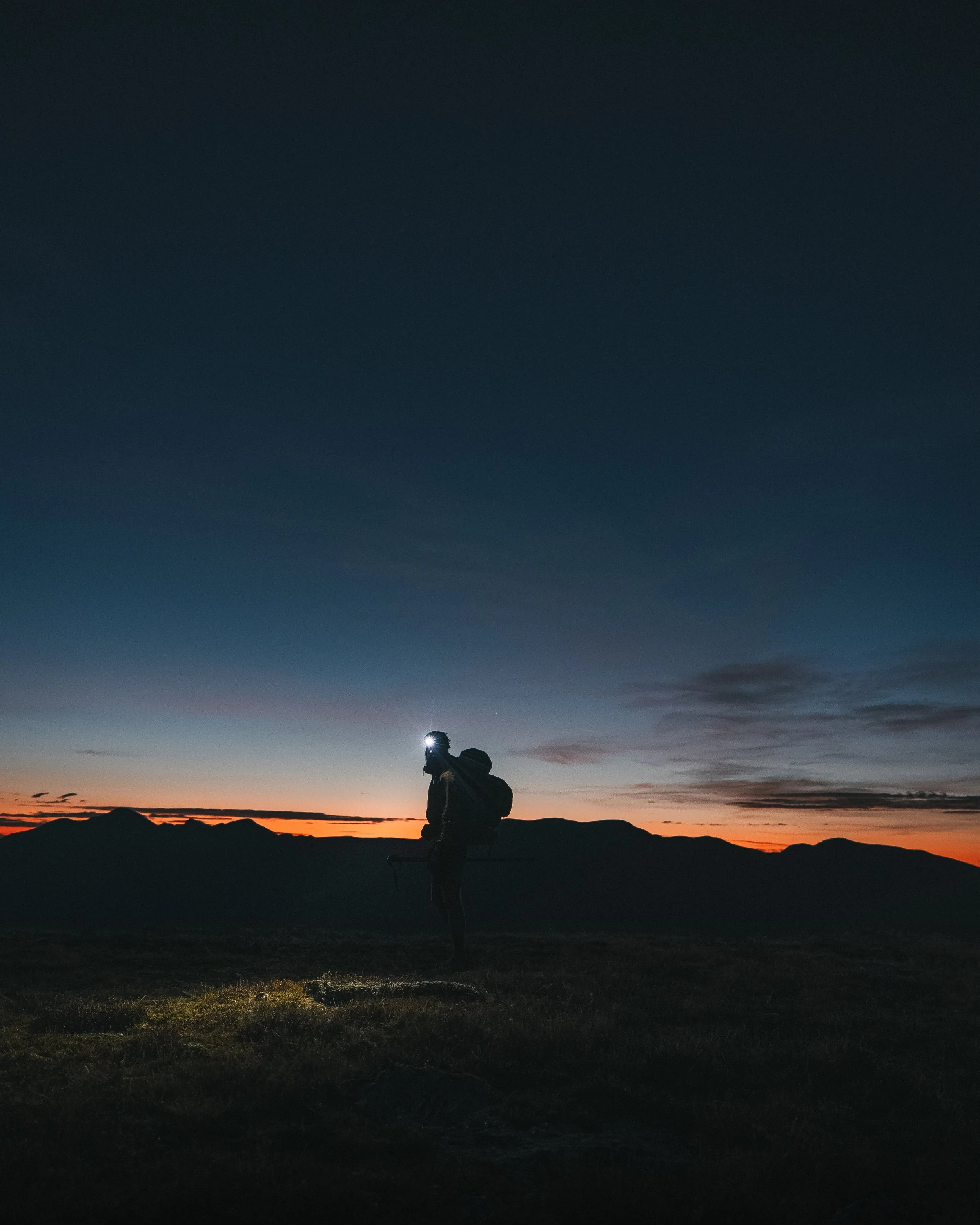 Silhouette of a person with a backpack and headlamp walking in a mountainous landscape at dusk or dawn.