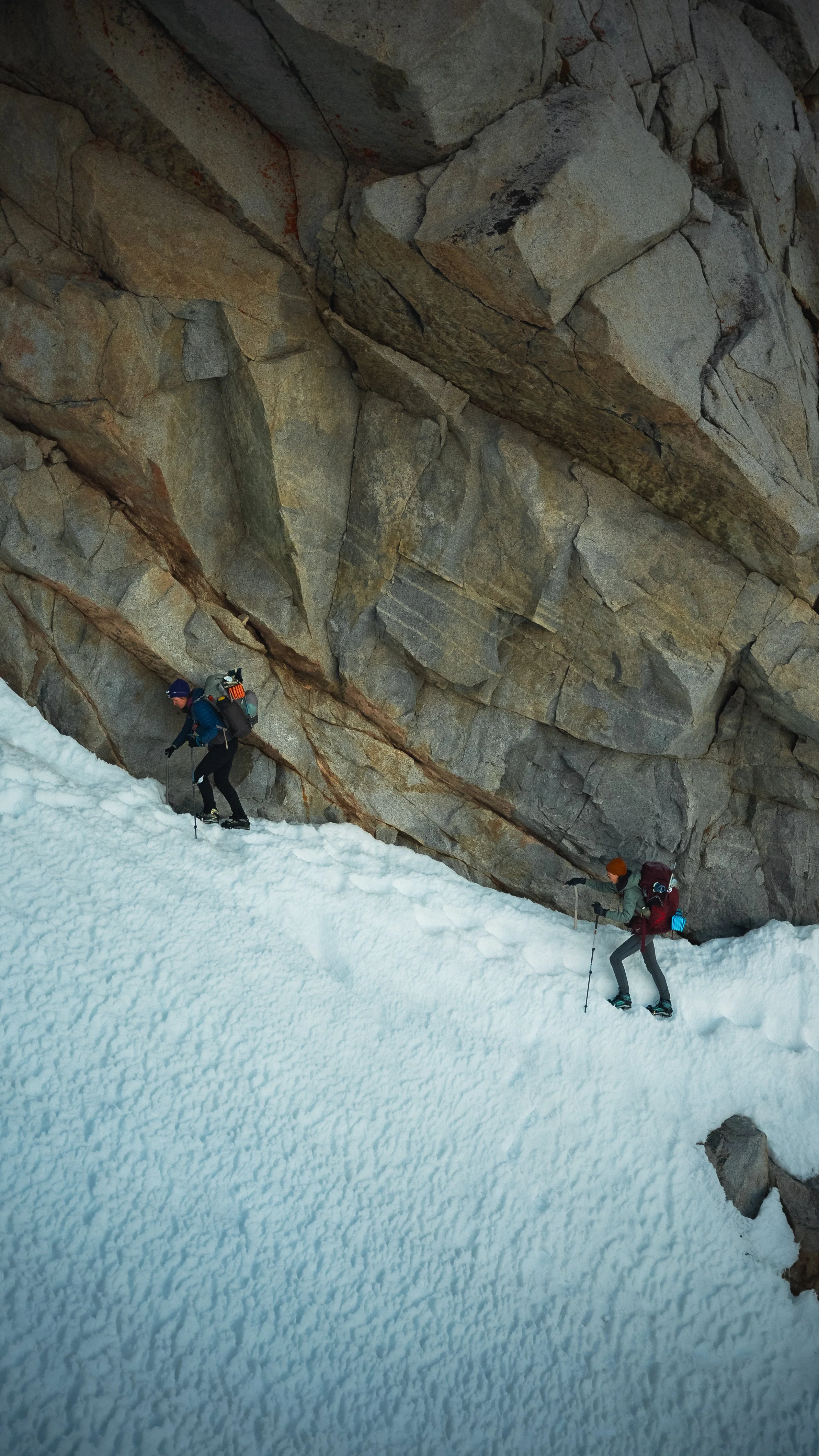 Two climbers ascending a snowy slope beneath a steep rocky cliff.