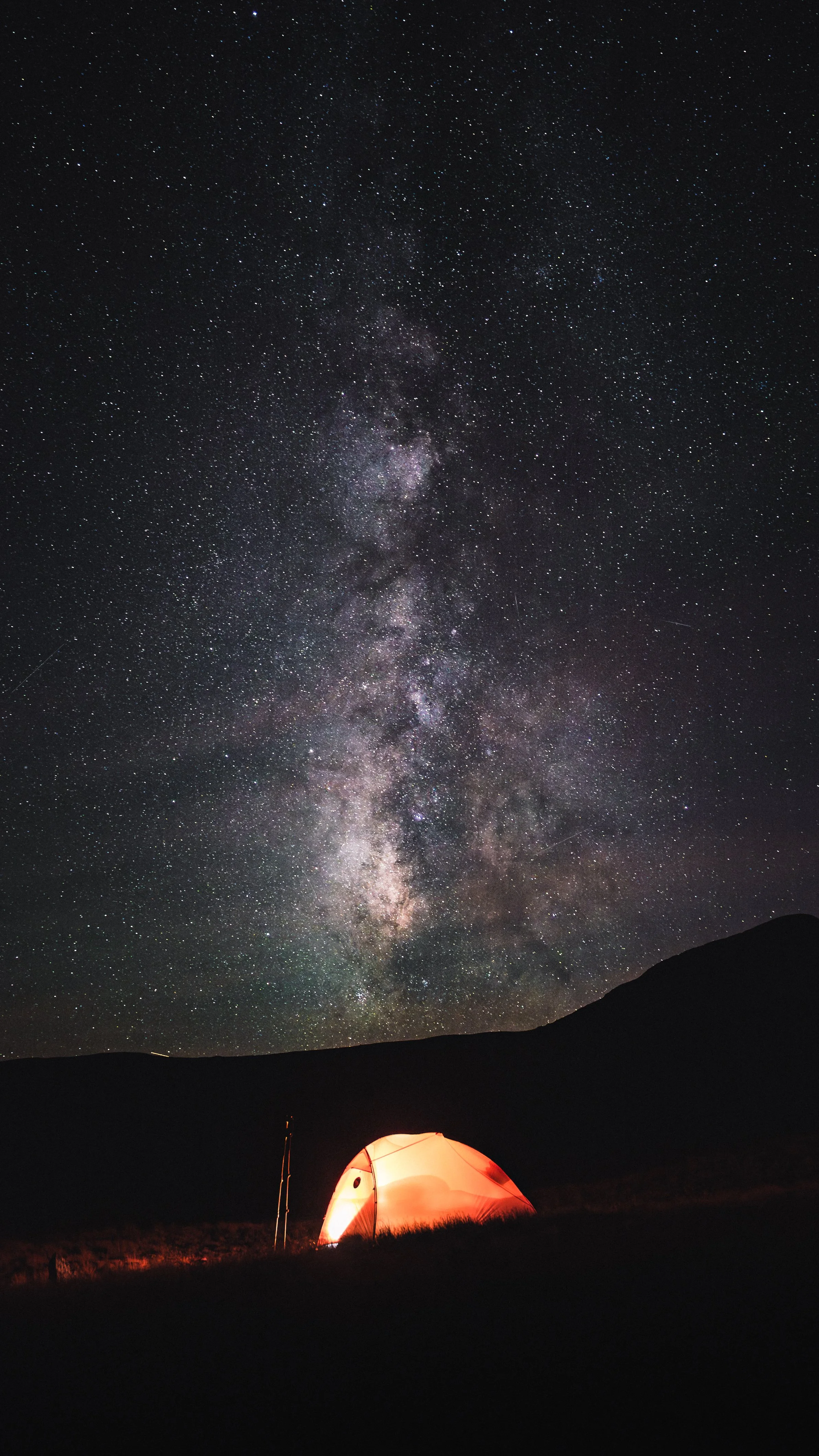 Night sky filled with stars and the Milky Way galaxy, with a glowing orange tent on the ground at the bottom.