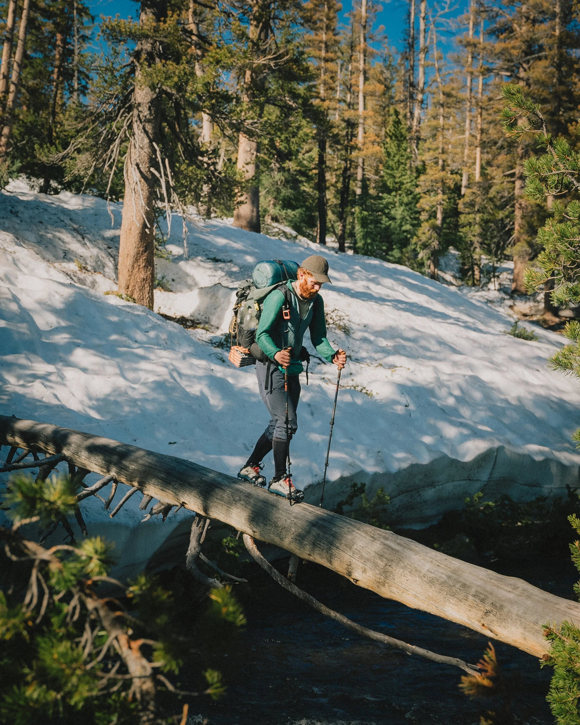 A man hiking in a snowy forest with trees, wearing a green jacket, black pants, hat, and carrying a large backpack, crossing a fallen tree over a stream.