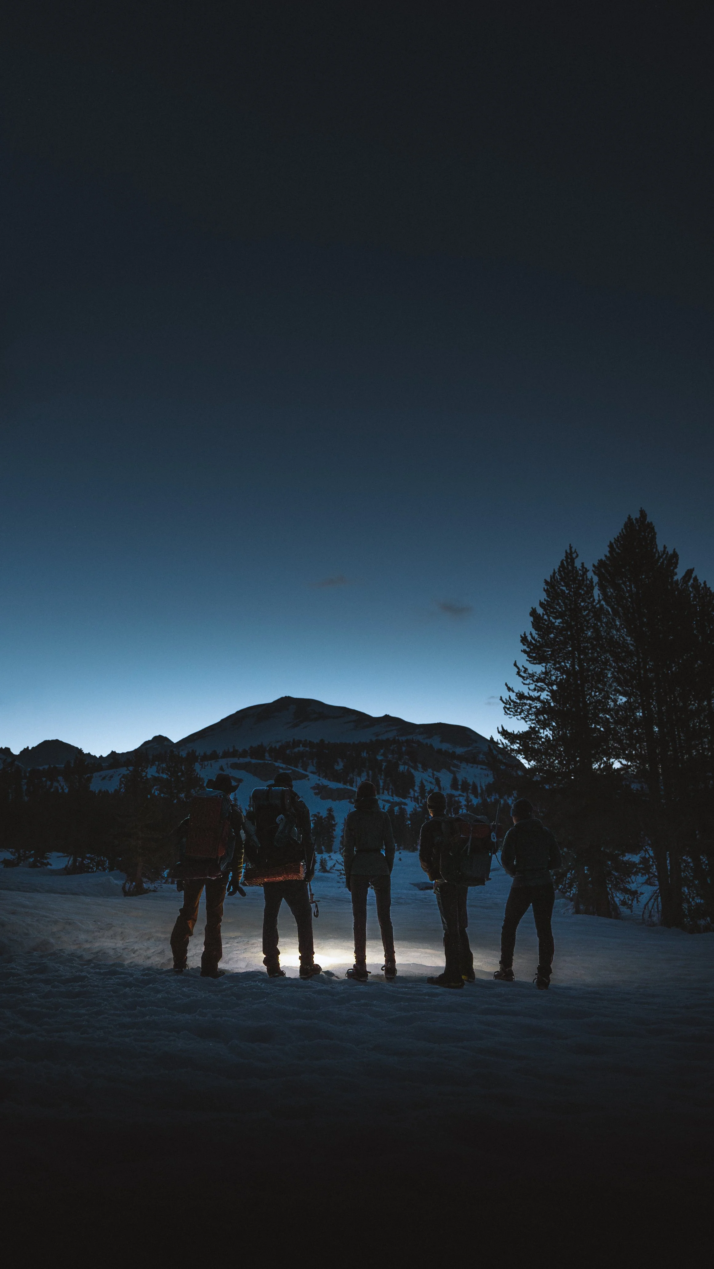Five hikers with backpacks and headlamps standing together on snowy ground at dusk, facing a mountain in the distance with a darkening sky.