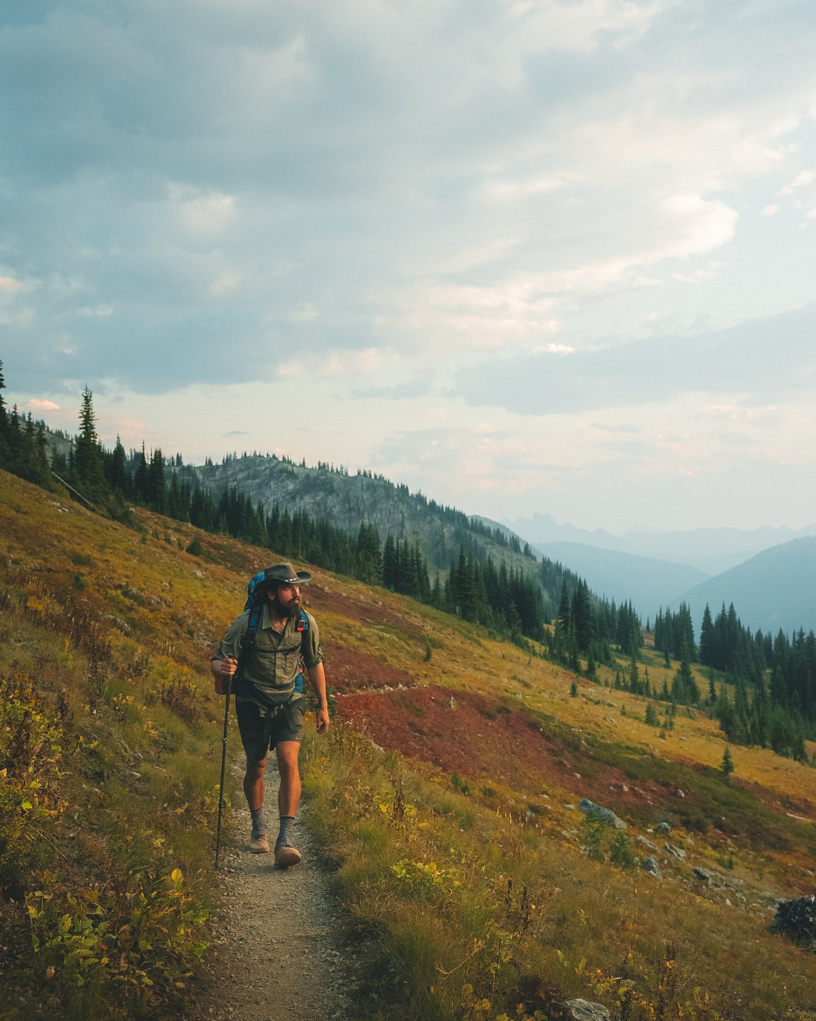 A man hiking with a backpack and trekking poles on a dirt trail through a mountainous landscape with trees and rolling hills under a cloudy sky.
