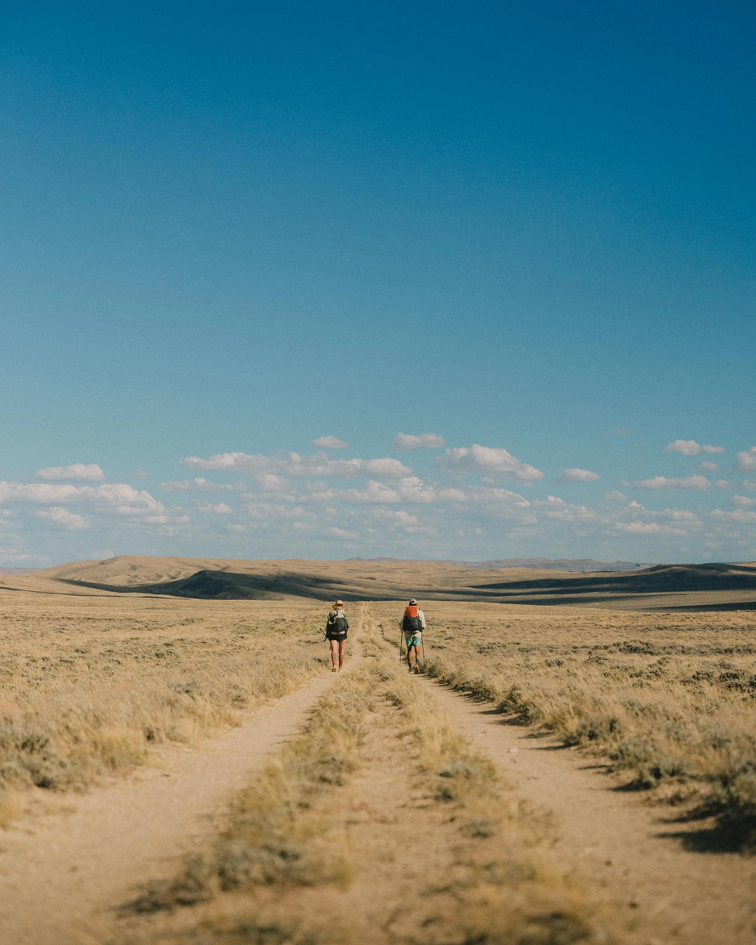 Two hikers walking down a dirt trail through a flat, grassy landscape under a bright blue sky with some scattered clouds.