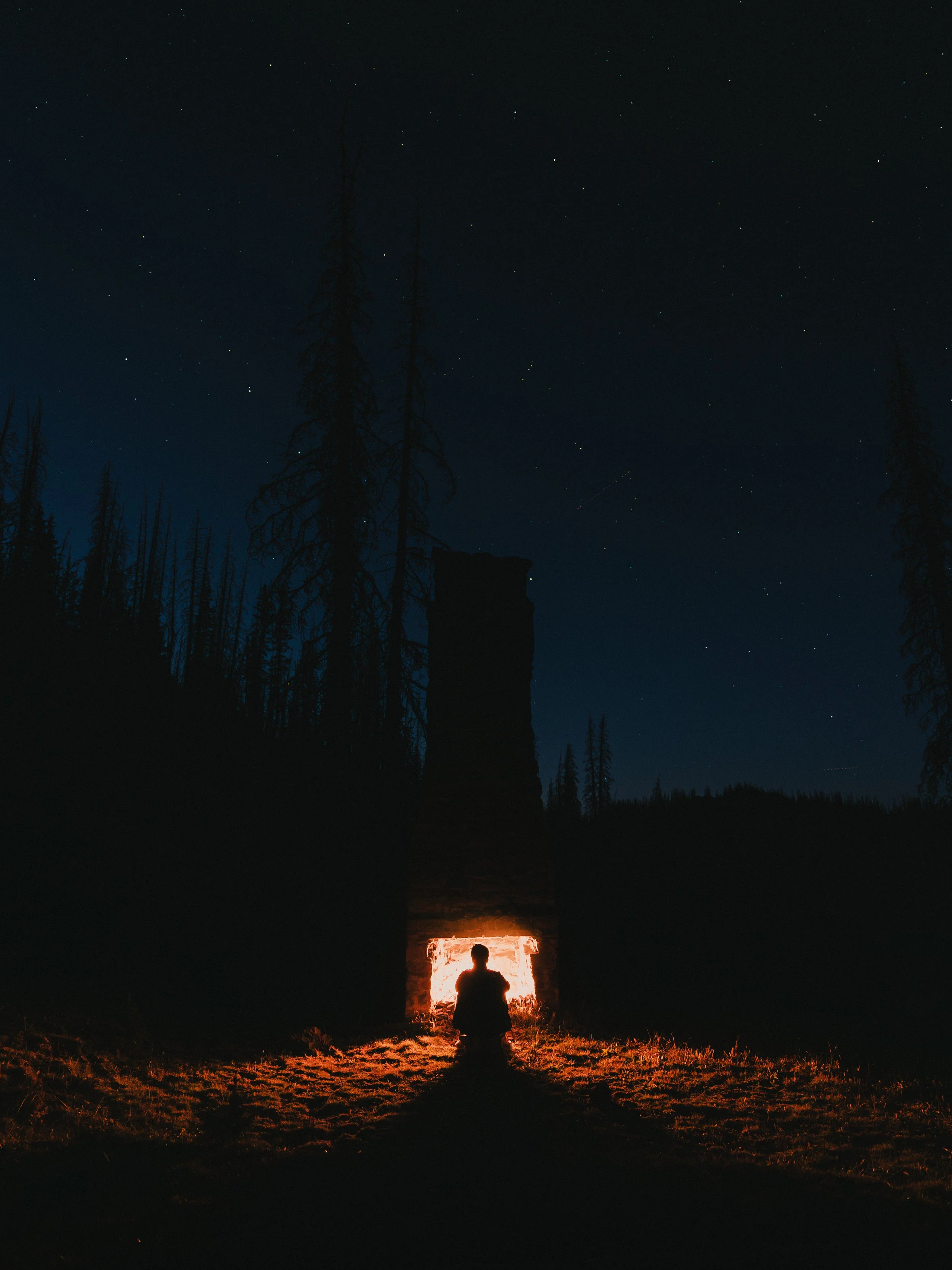 Person sitting on the ground by a campfire at night, with a large brick chimney structure behind them and a starry sky above, surrounded by tall trees.