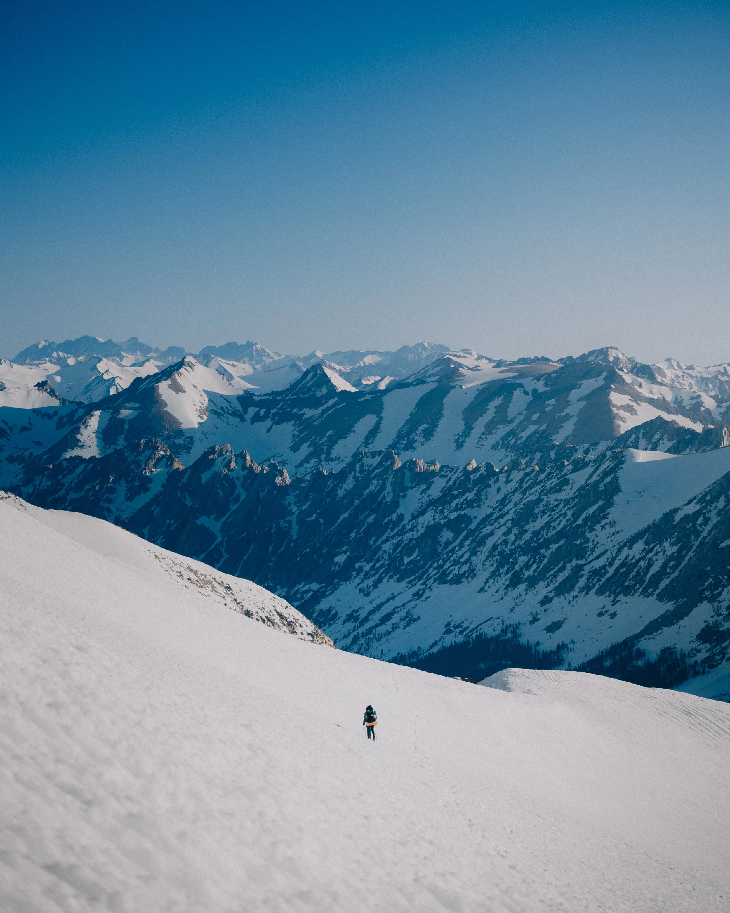 Solo hiker walking through snow-covered mountain landscape with rugged peaks in the distance and clear blue sky.