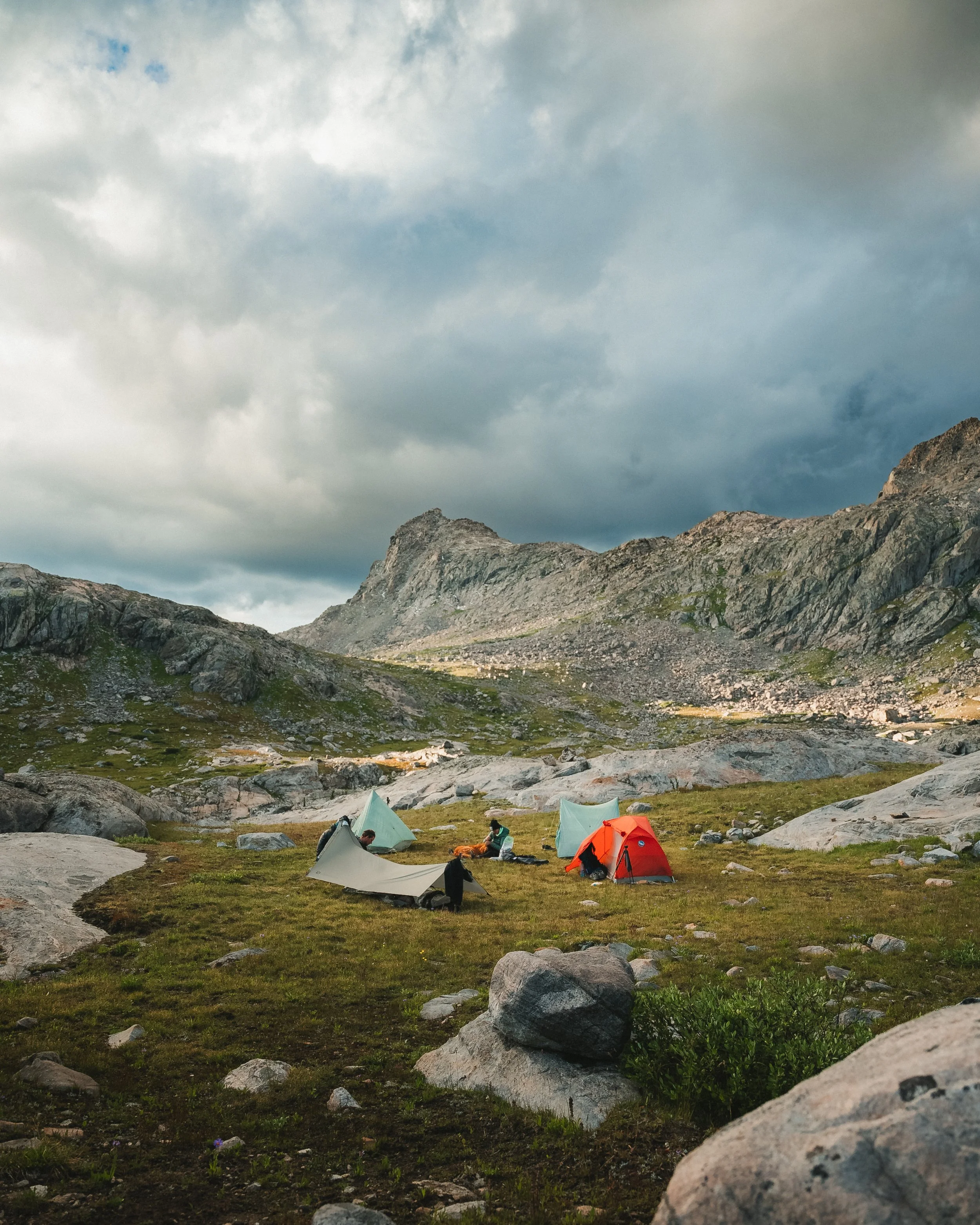 People camping in tents on a grassy patch with rocks, mountain peaks in the background, under a cloudy sky.