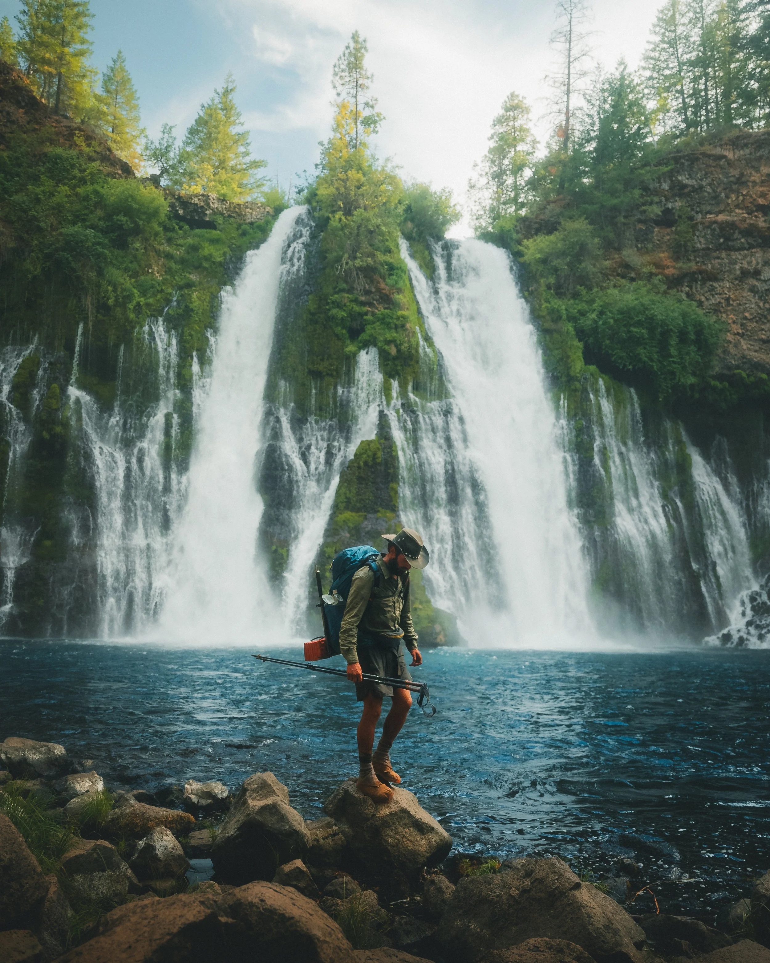 Hiker with a trekking pole and backpack standing on rocks at the edge of a lake in front of a large waterfall surrounded by green trees.