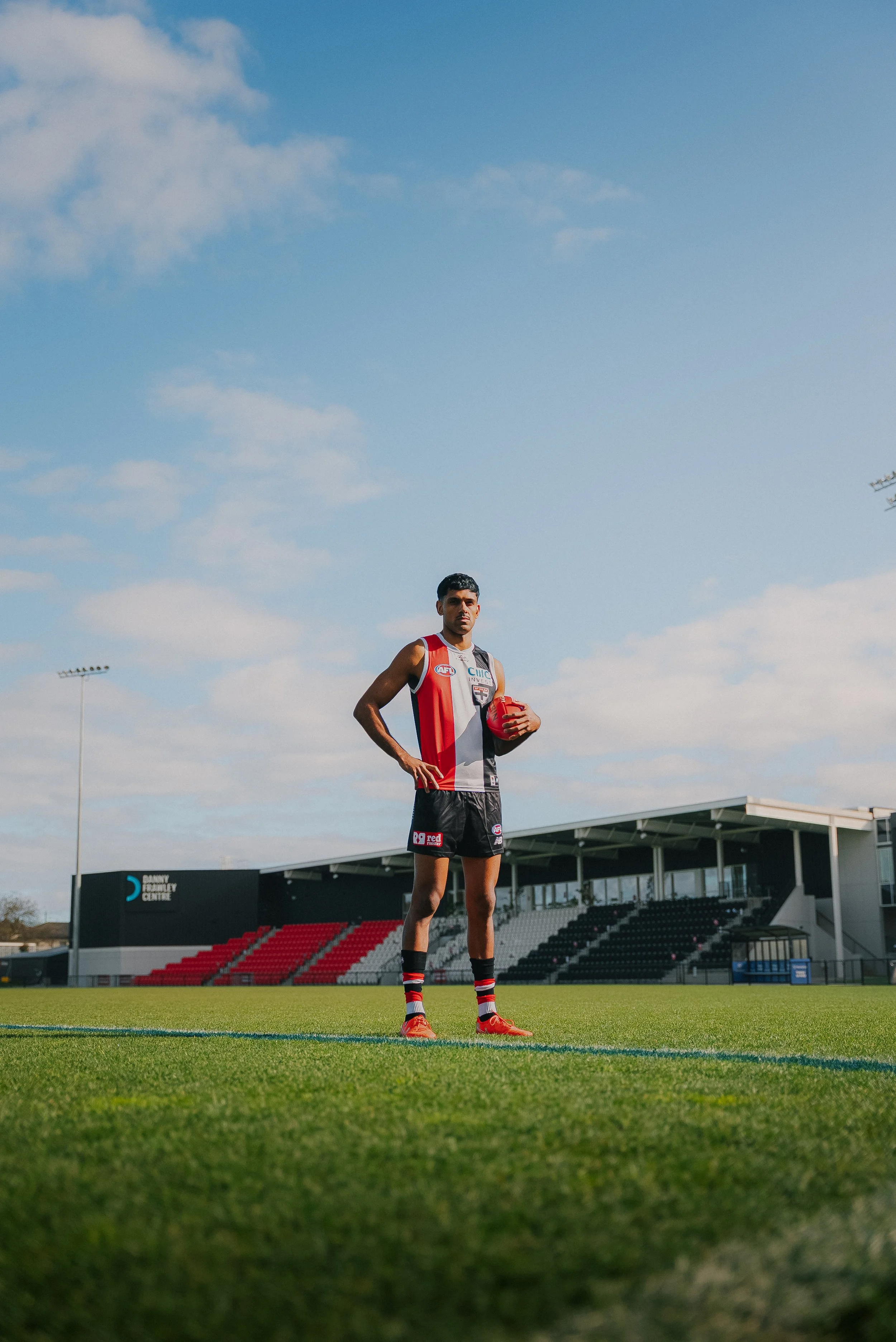 An Australian football player stands on a grass field holding a red football, dressed in team uniform with black shorts, red shoes, and striped socks, with a stadium in the background under a partly cloudy sky.