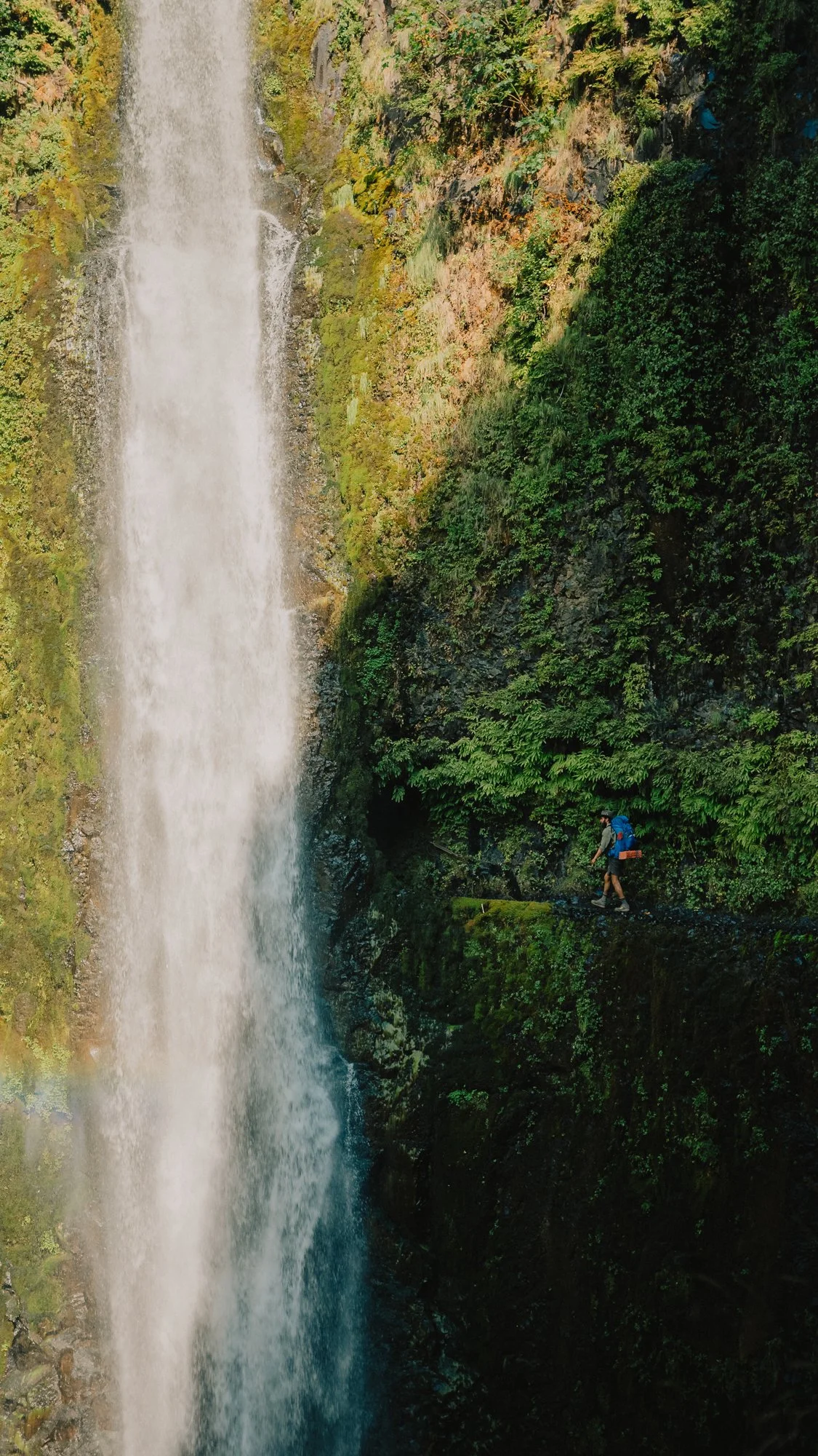 Hiker with backpack walking along a narrow trail next to a tall waterfall in a lush green forest.