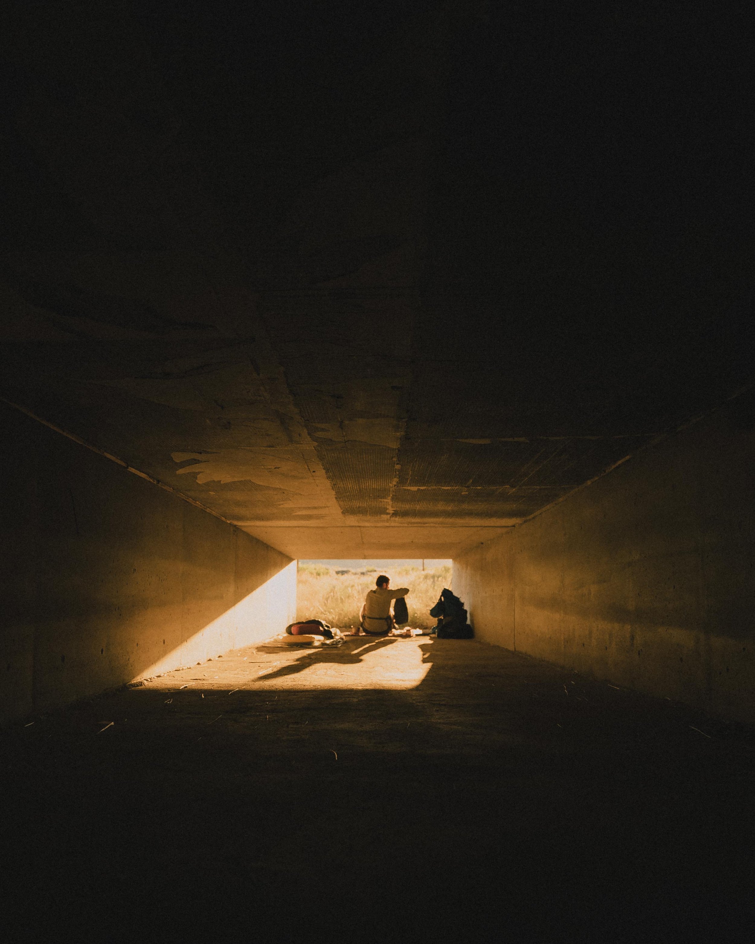 Person sitting inside a tunnel with sunlight shining in, sitting next to backpacks, facing outward toward an open field.