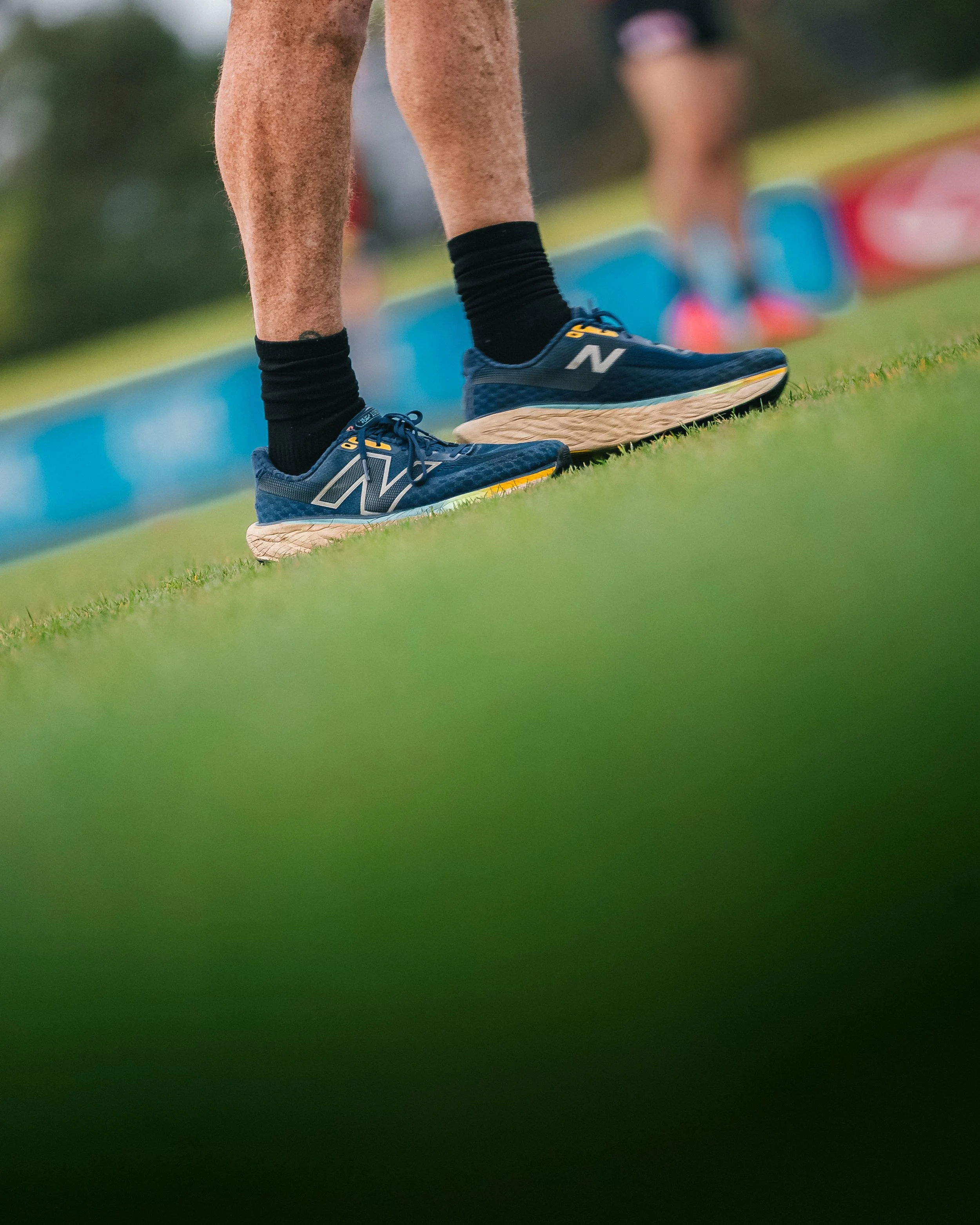 Close-up of a person wearing blue New Balance running shoes and black socks, standing on a grass field with blurred people in the background.