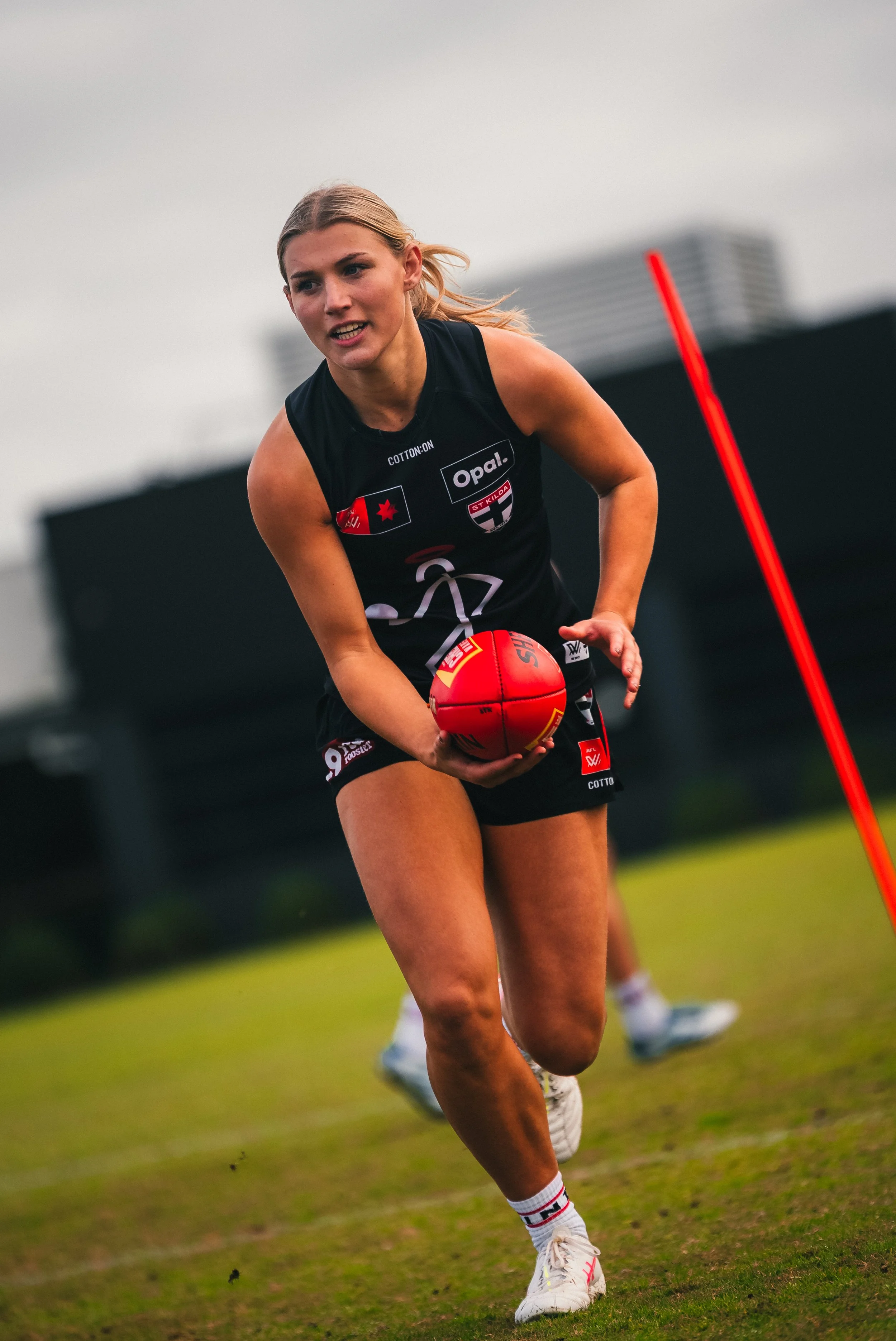 An female athlete running on a sports field while holding a red football, wearing a black sports uniform with various logos, in an outdoor setting with a cloudy sky and a partially visible building in the background.