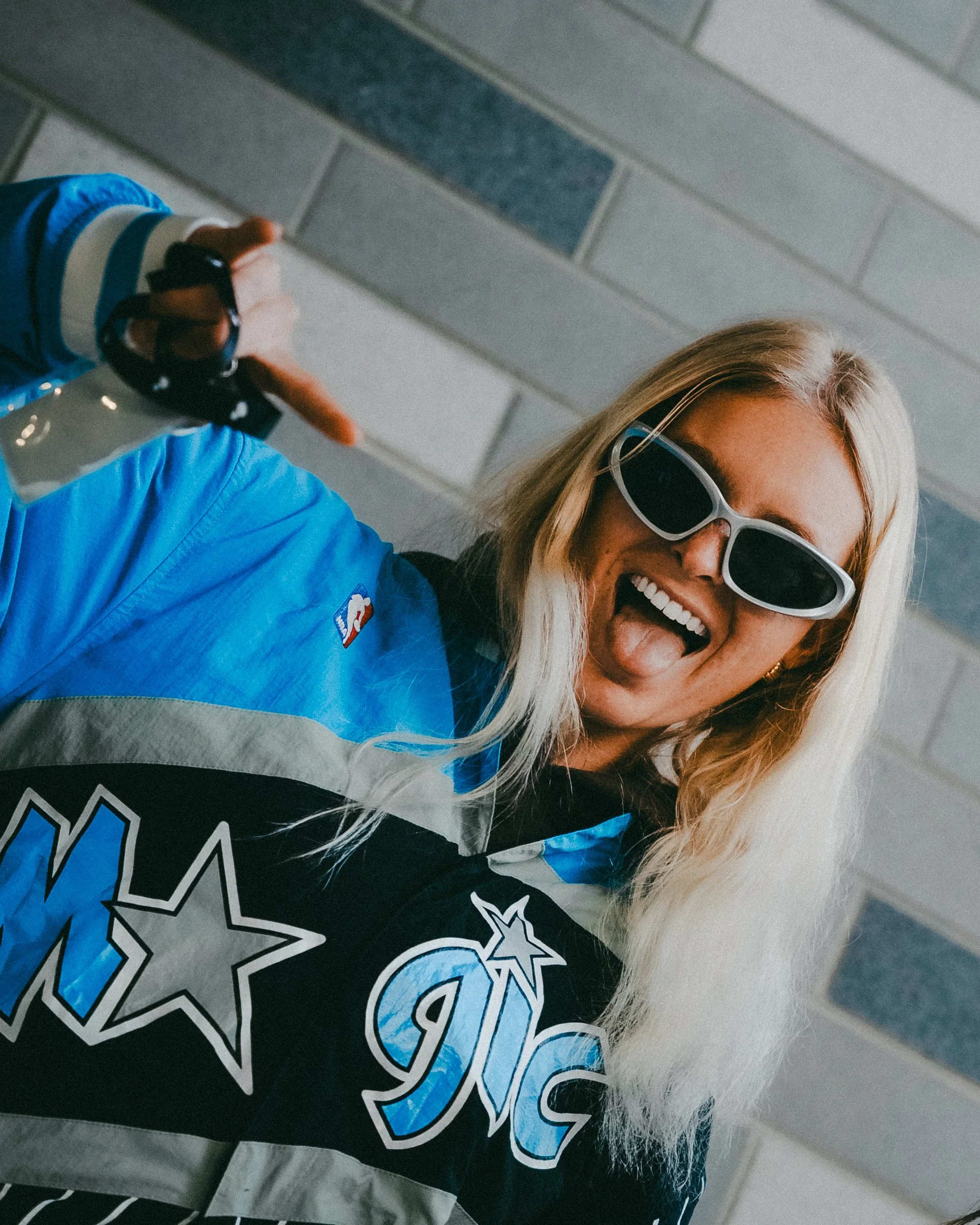 Woman wearing sunglasses and a blue and black sports jacket, smiling with tongue out, holding a water bottle, standing against a brick wall.