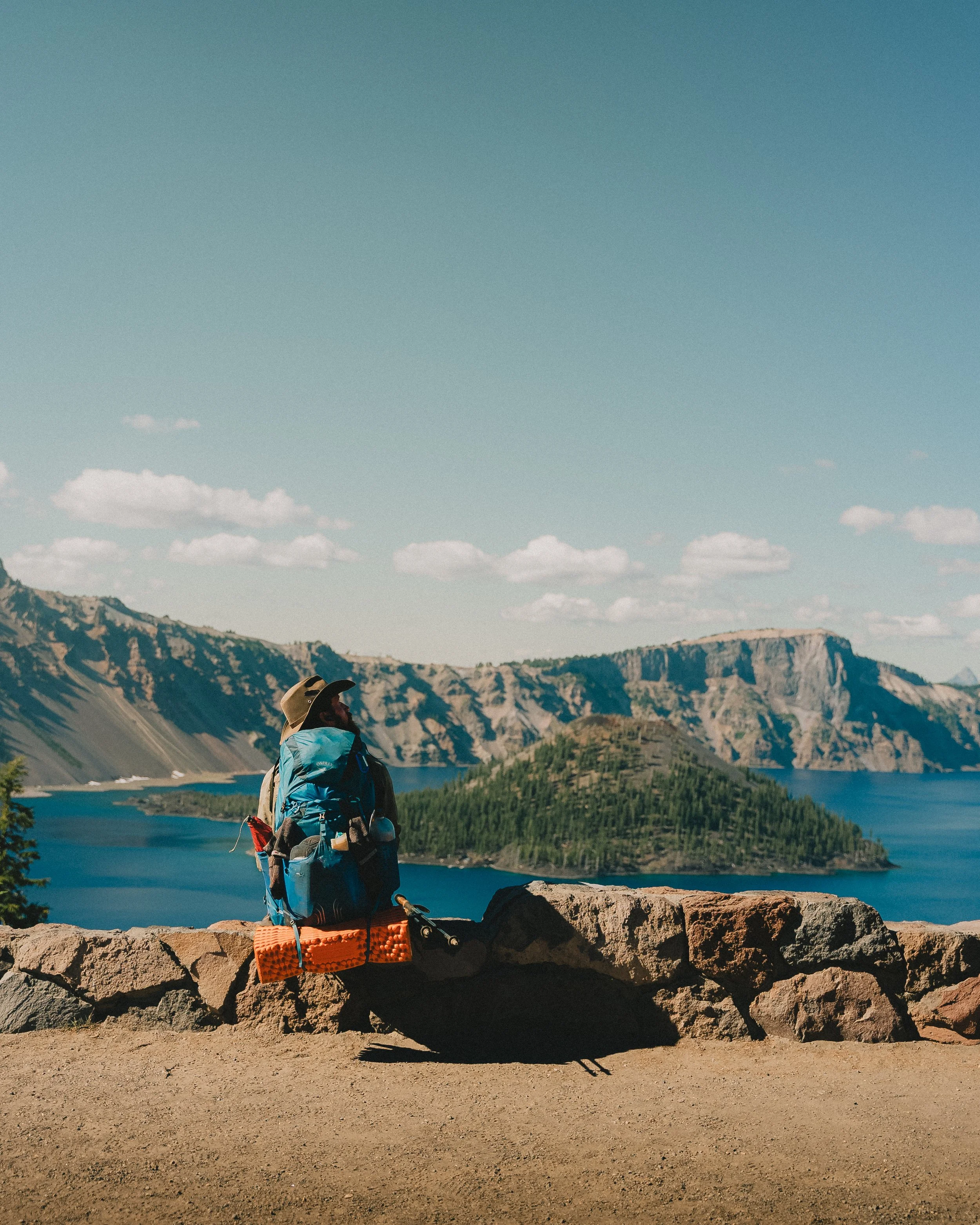 A hiker with a large blue backpack sitting on a stone wall overlooking a scenic lake with a forested island, mountains, and a blue sky with clouds.