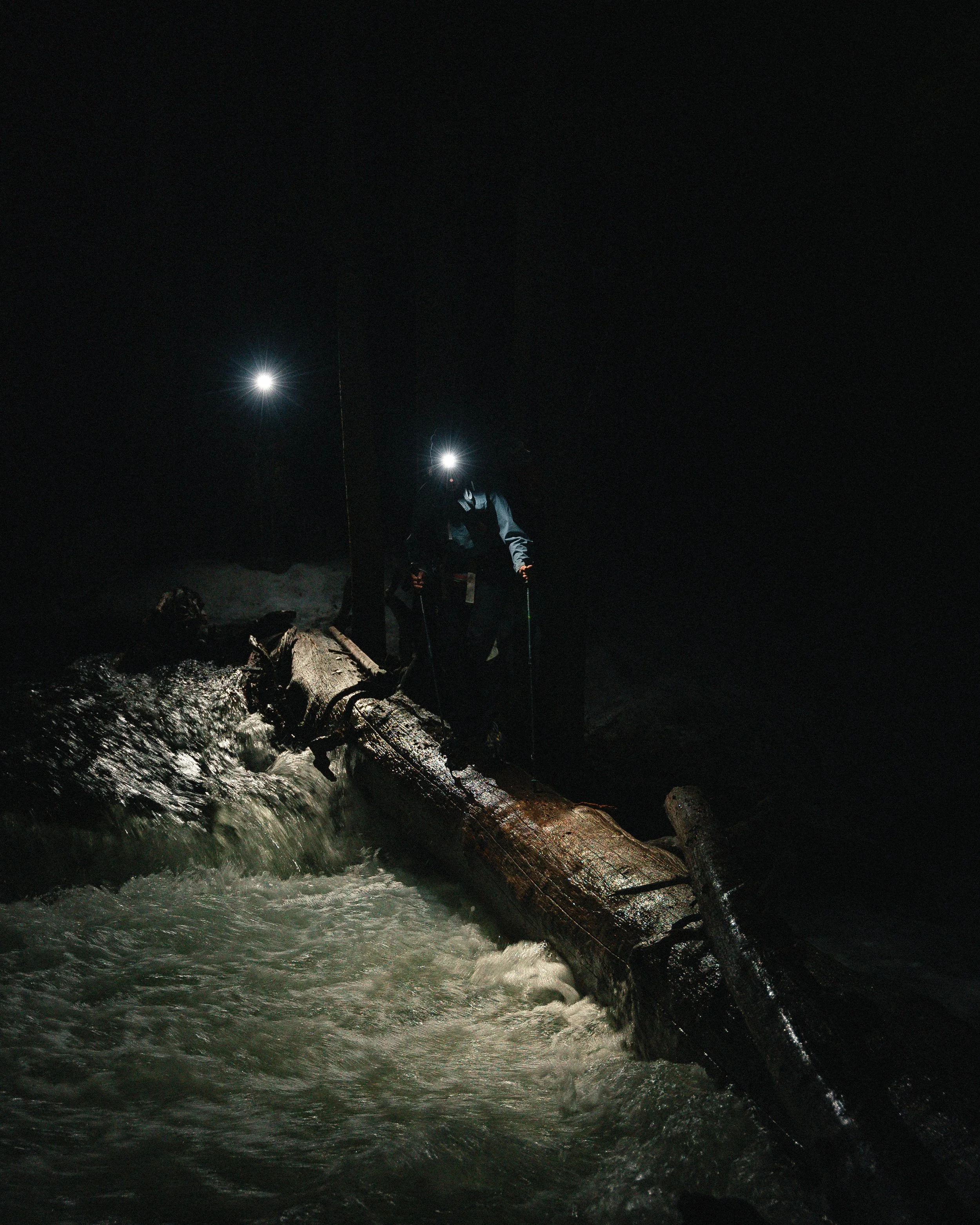 Person with headlamp standing on a fallen tree across a rushing river at night.