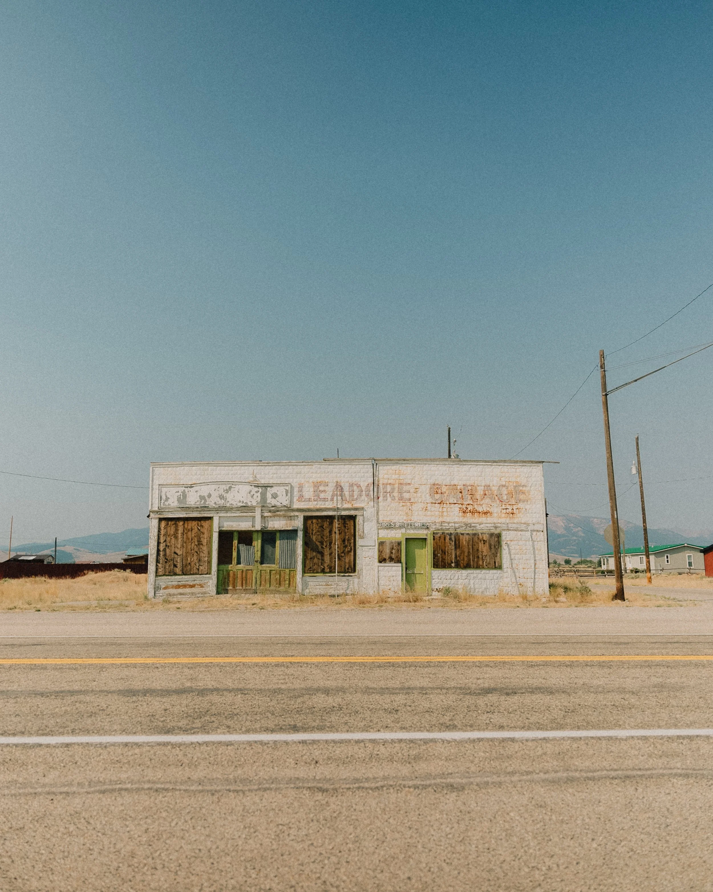 A weathered, abandoned building in a rural area under a clear blue sky, with some distant mountains in the background and power lines in front.