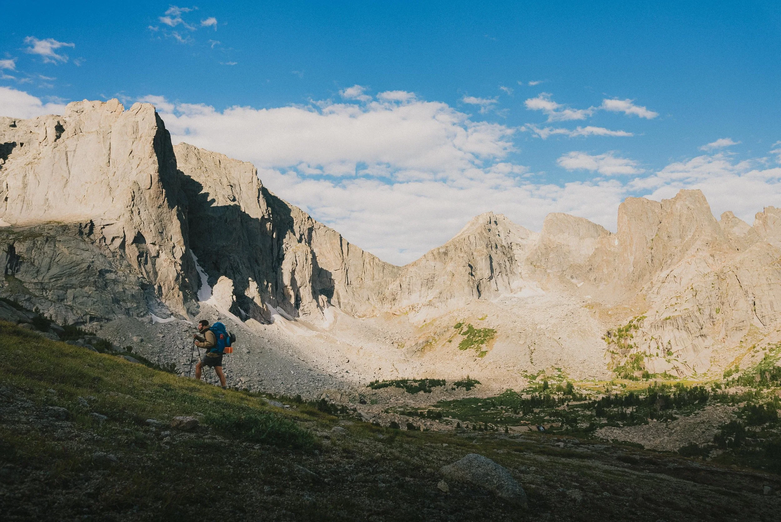 A hiker walking in a mountainous landscape with towering rocky peaks and a partly cloudy sky.