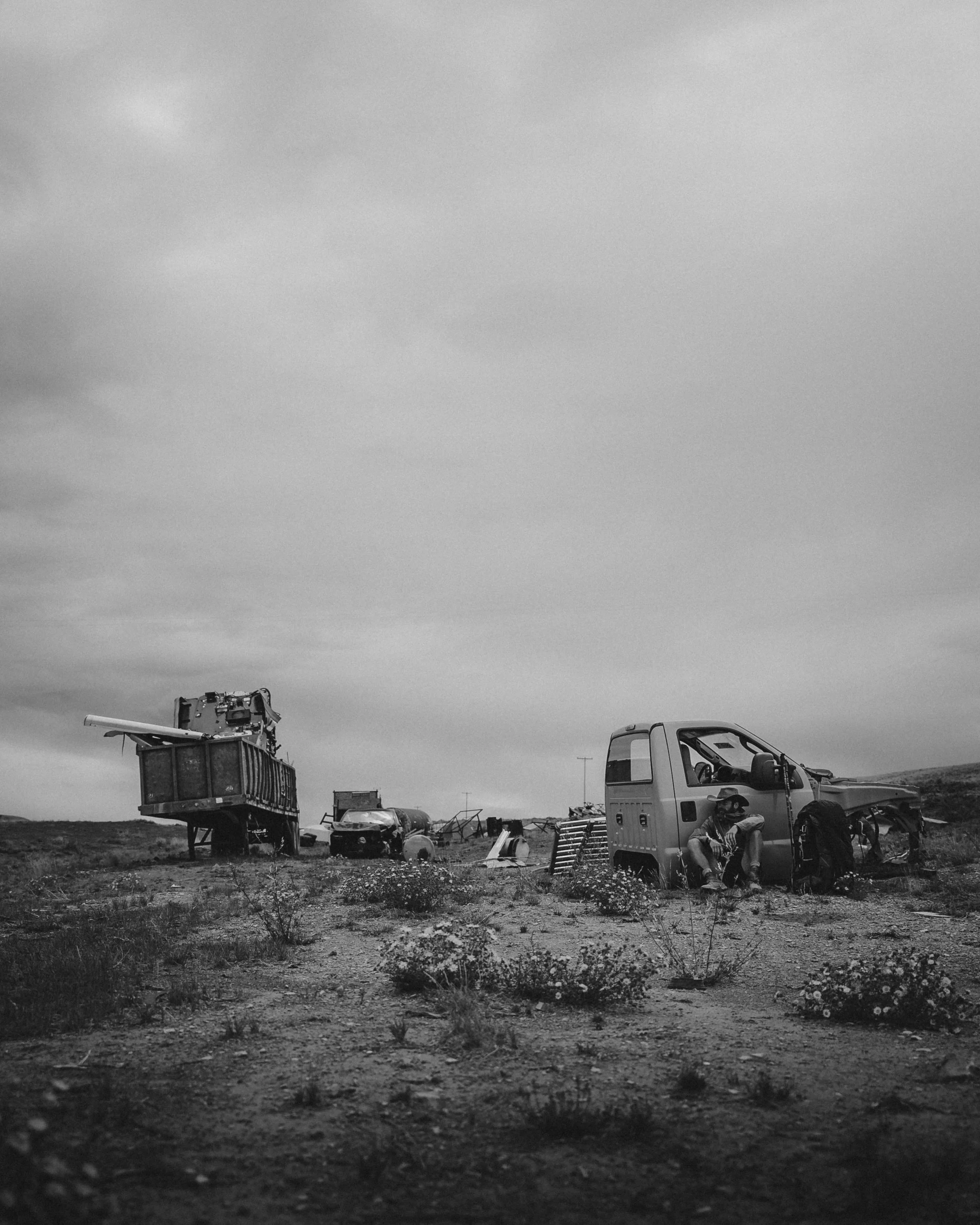 A black and white photograph of a desert landscape with abandoned vehicles and a person sitting on the ground near a small truck.