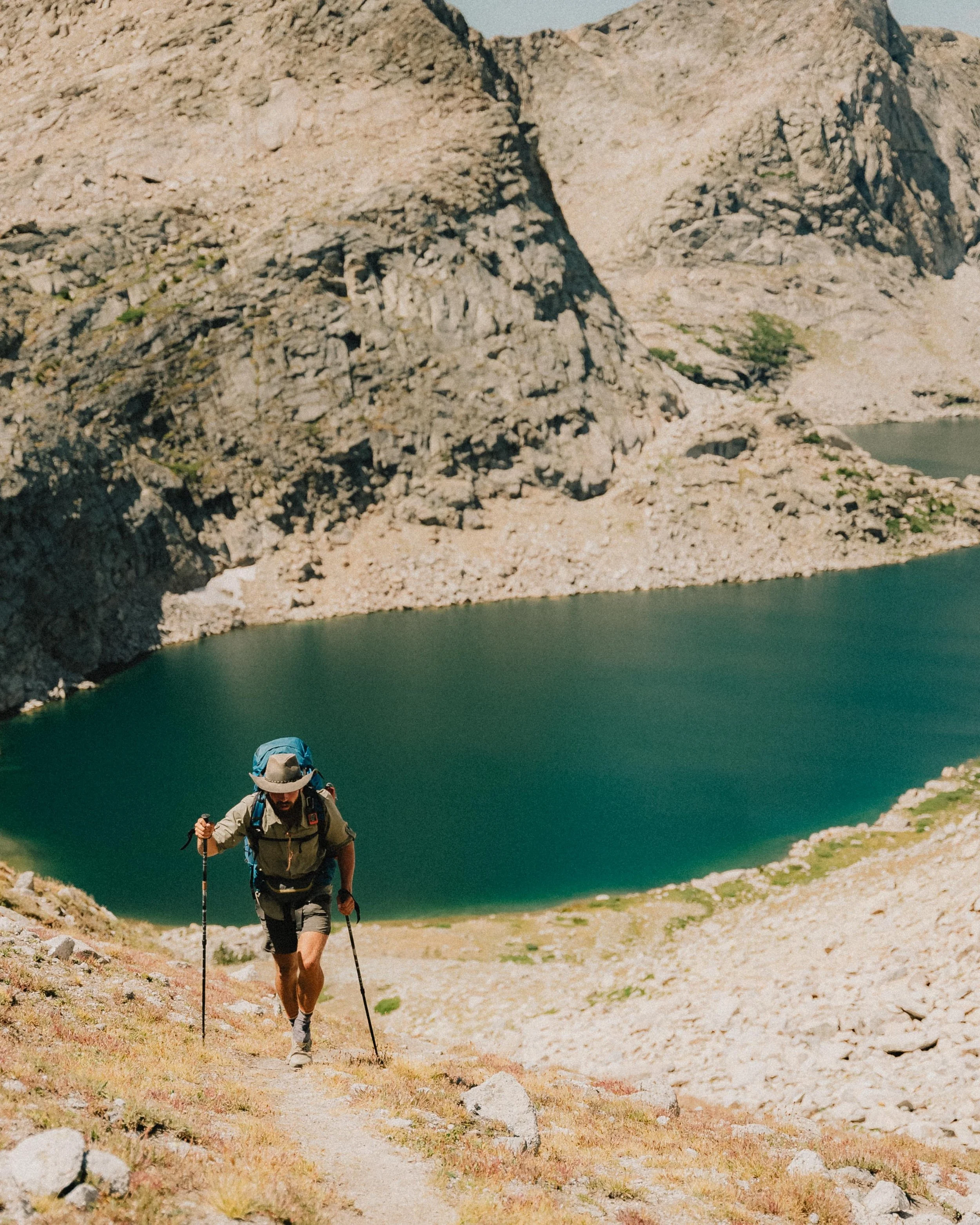 A man hiking up a mountain trail near a lake with steep rocky cliffs in the background.