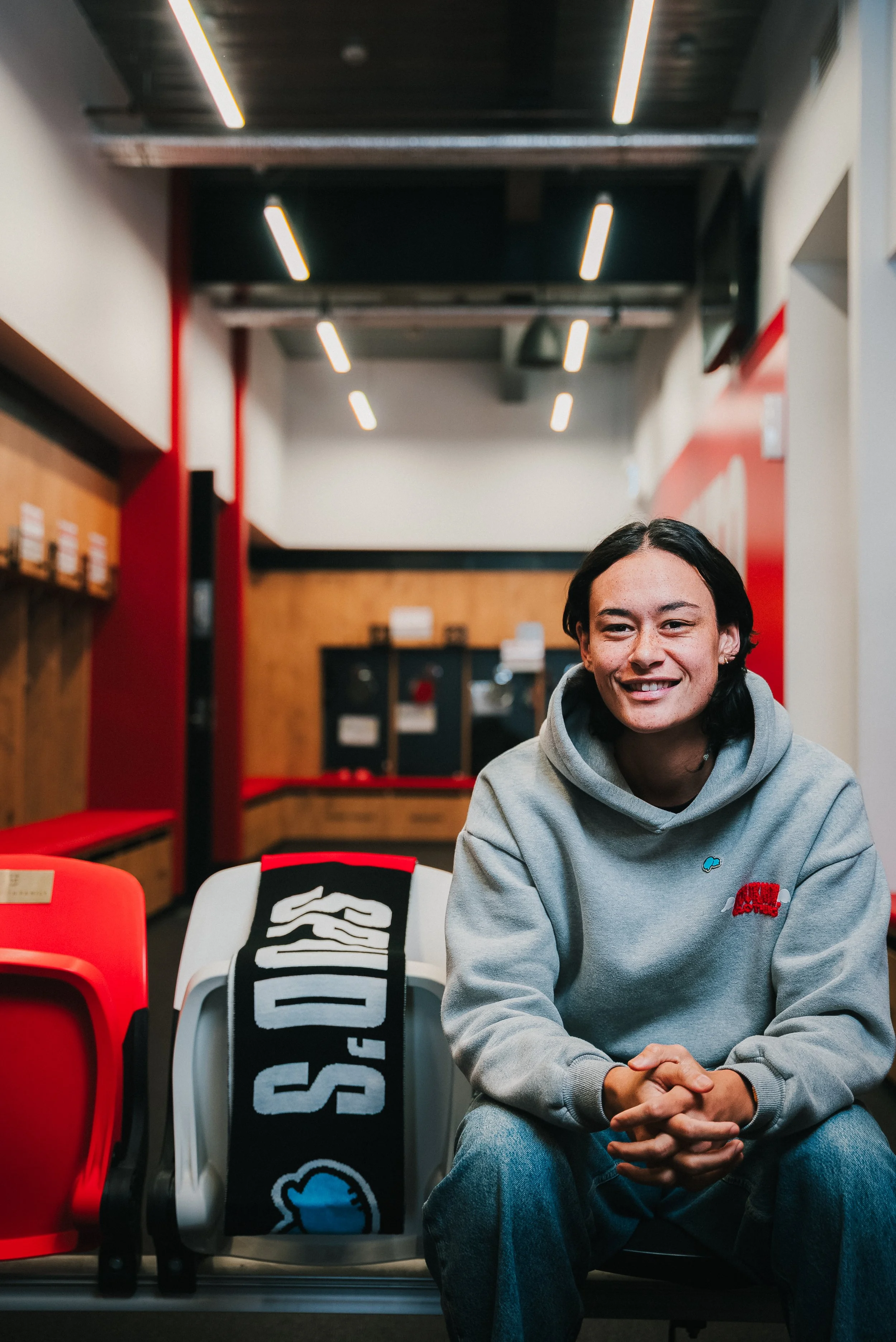 A young man with dark hair, wearing a gray hoodie, sits smiling in a modern, brightly lit indoor space with red and wood decor.