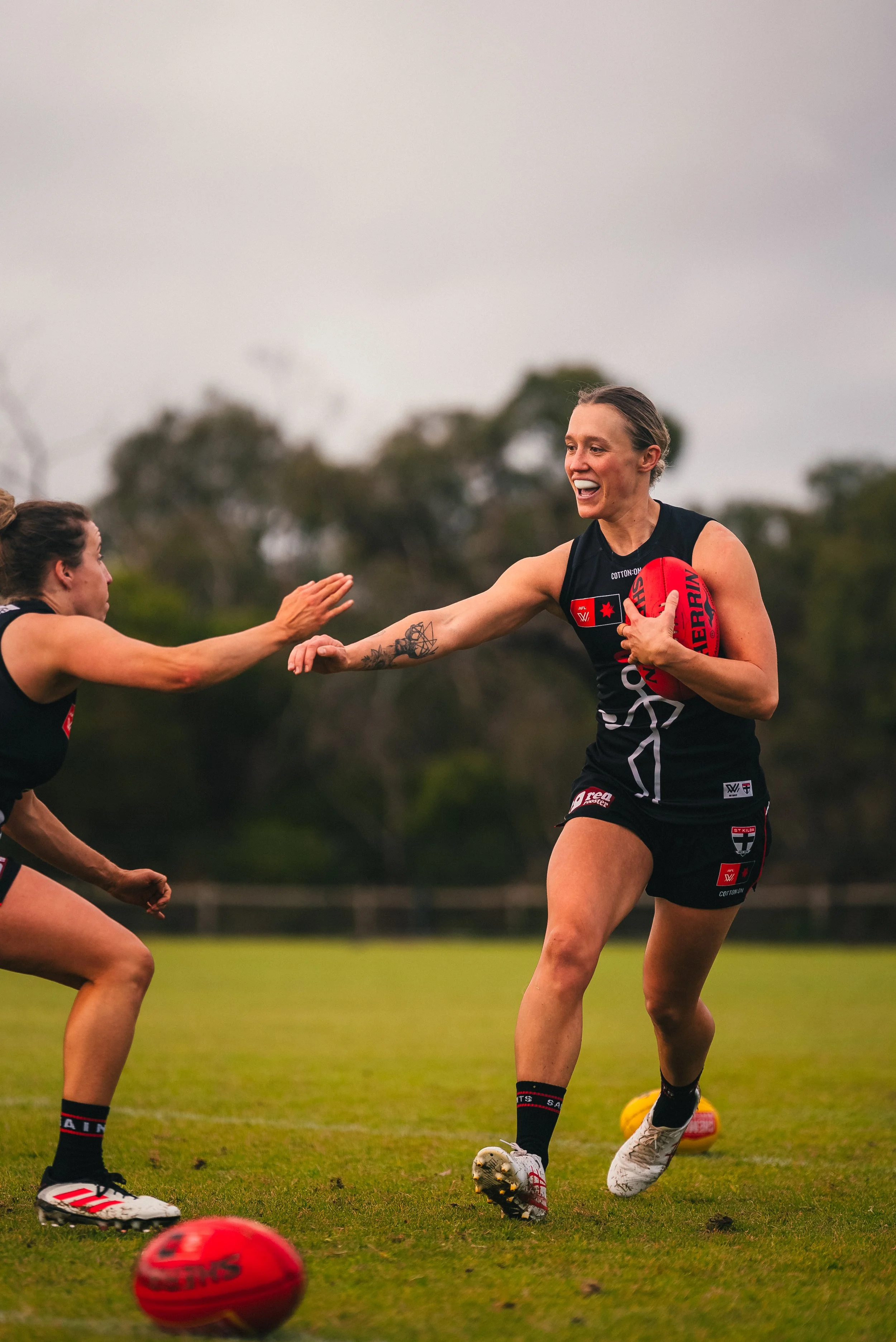 Two female athletes playing Australian rules football on a grassy field, one with a red football and the other reaching out for a high five.