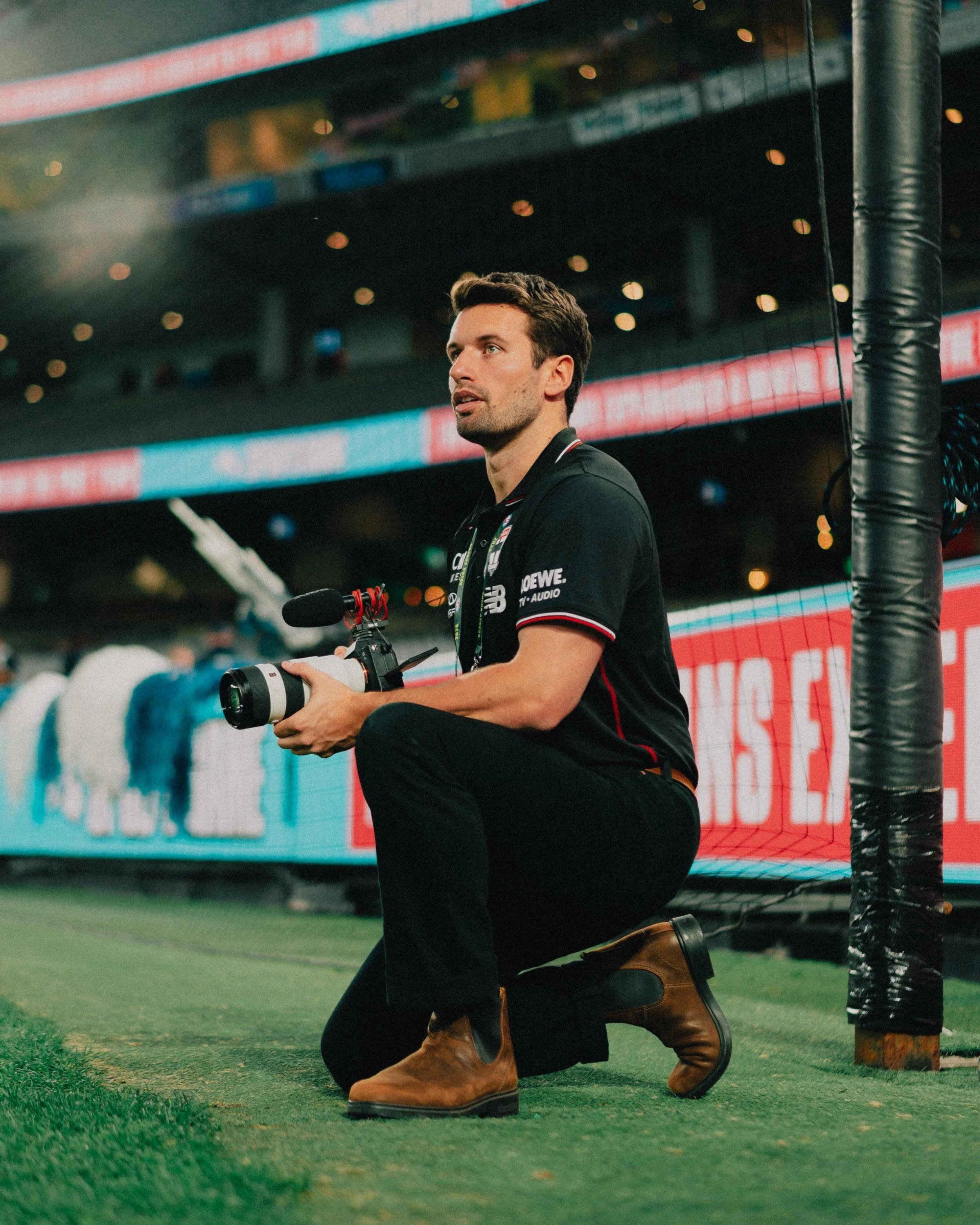 A man kneeling on a sports field holding a professional camera with a telephoto lens, wearing a black polo shirt and brown boots.
