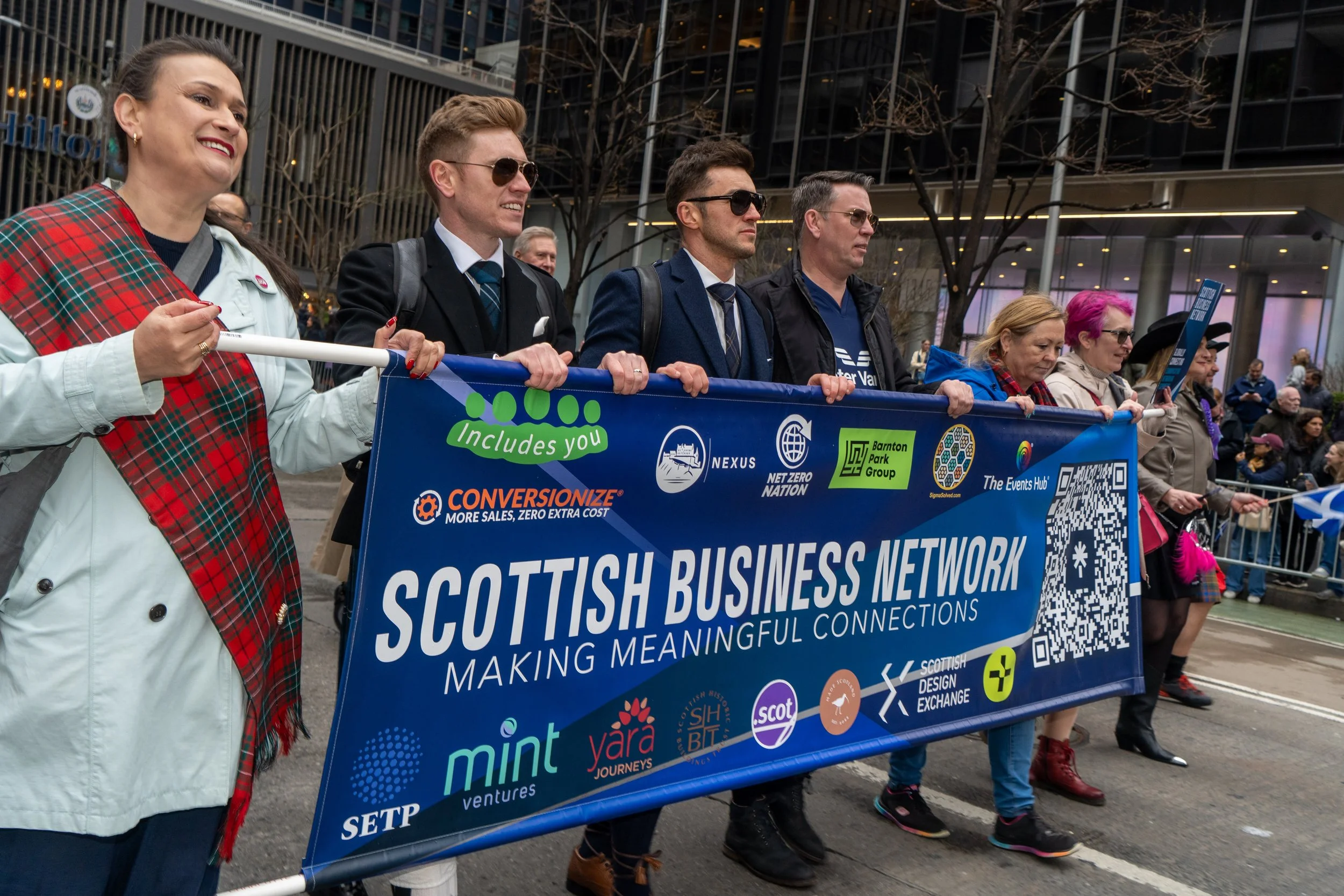 People participating in a parade march holding a blue banner that reads 'Scottish Business Network' with additional text and logos, in an urban setting with onlookers in the background.