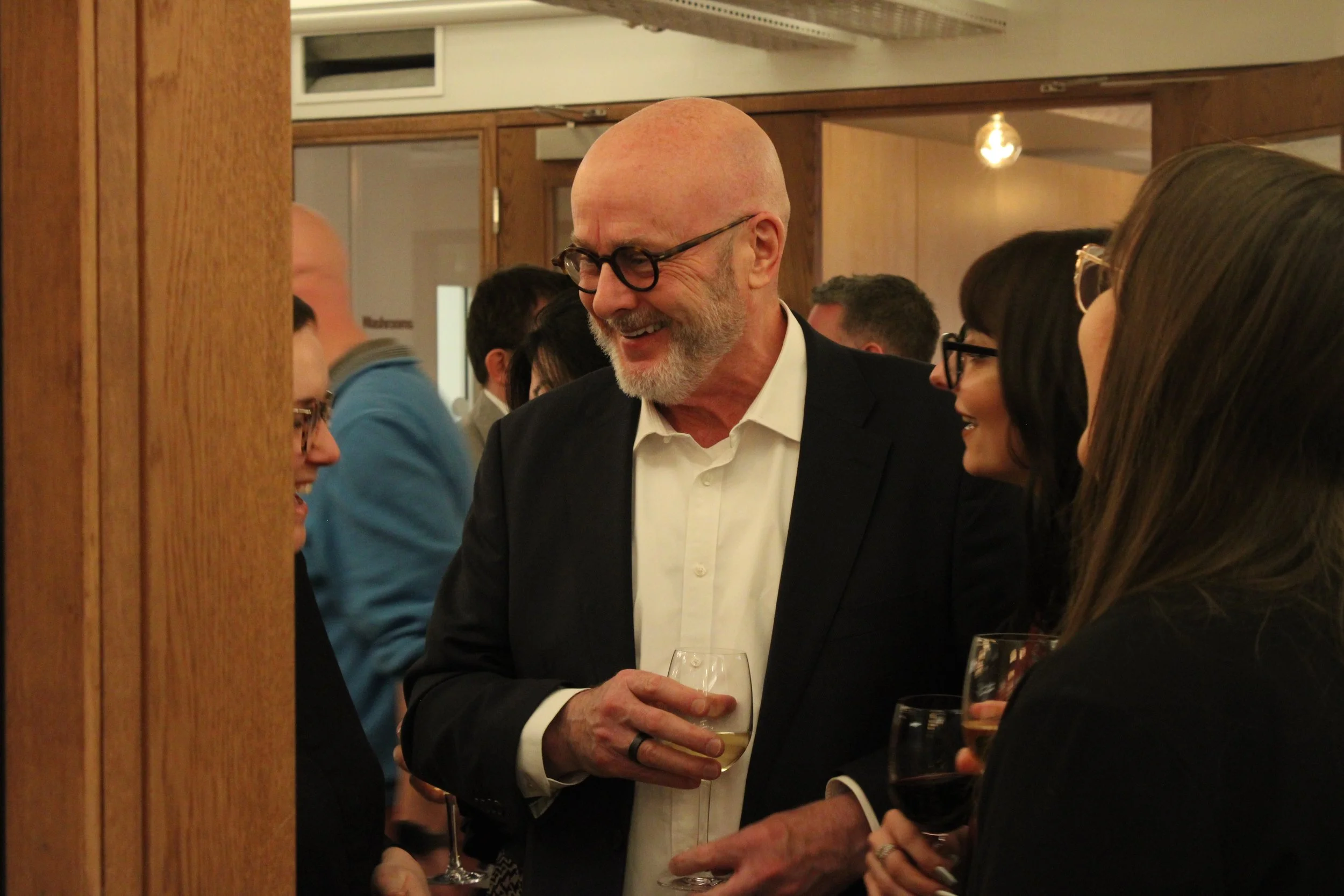 A group of people socializing at a gathering, with four individuals in foreground holding wine glasses, engaging in conversation and smiling in an indoor setting with warm lighting and wooden decor.