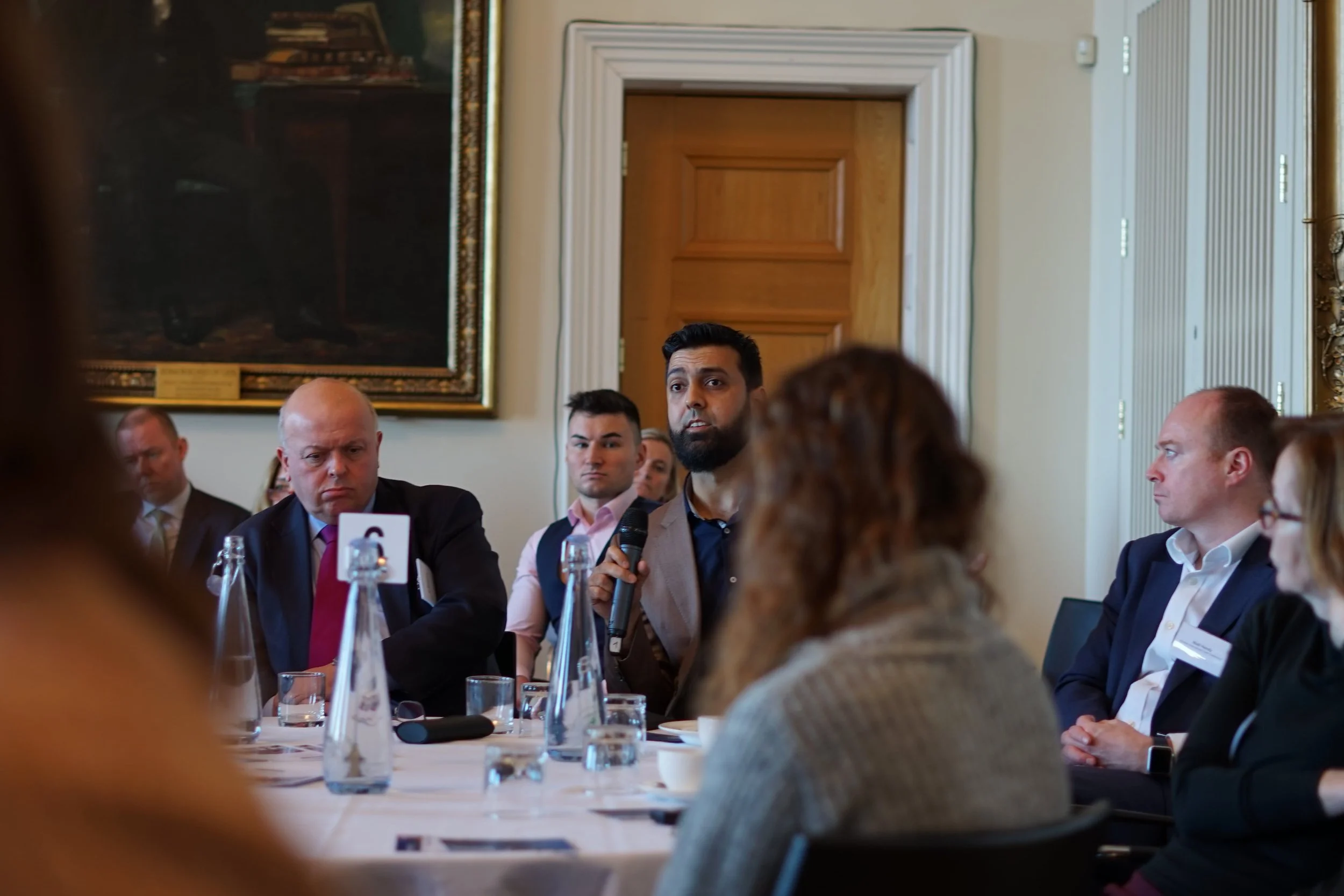 A man with a beard holding a microphone is speaking at a formal meeting or conference, seated at a round table with other attendees in a room with ornate woodwork and a large framed portrait on the wall.