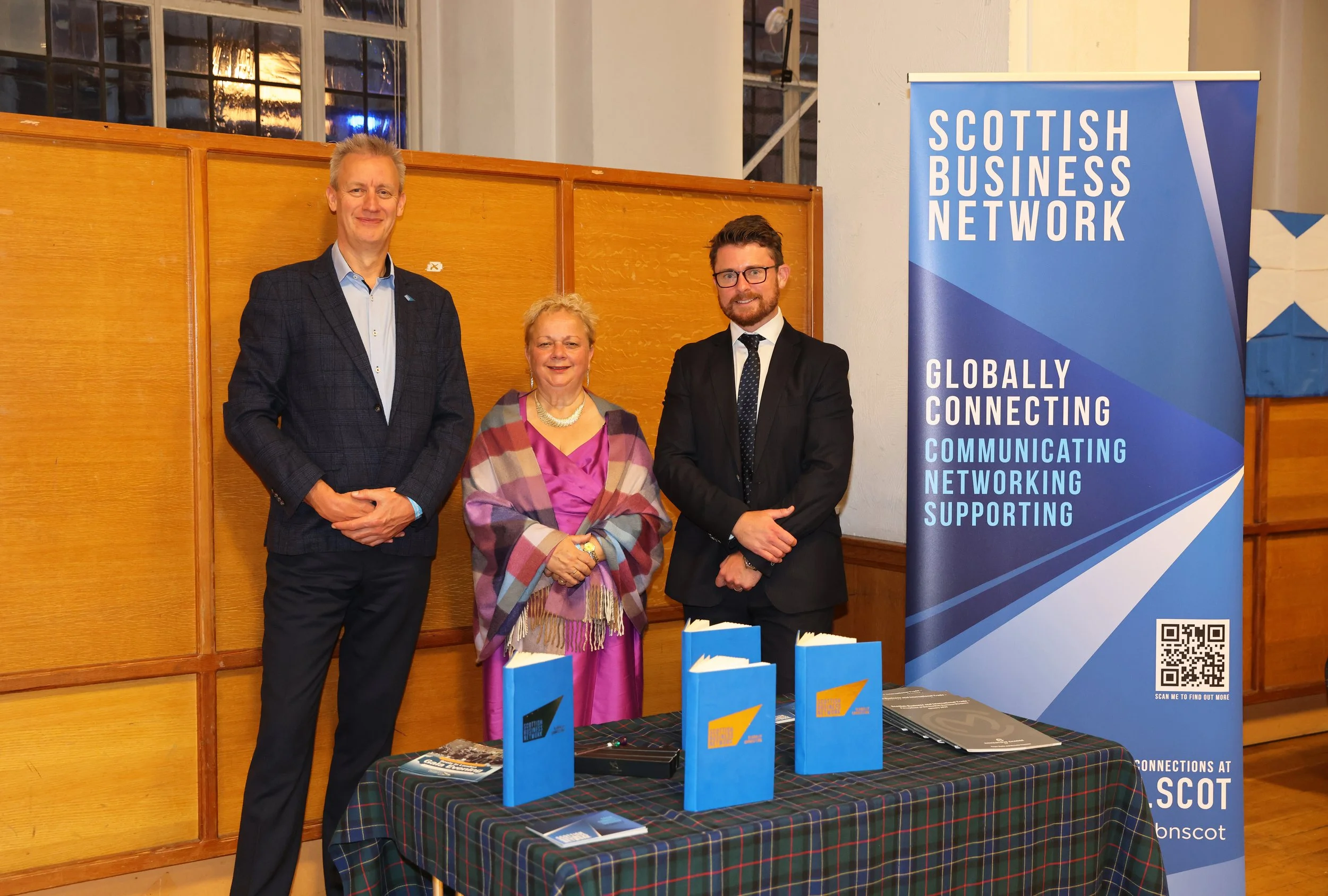 Three people standing behind a table at a Scottish Business Network event. The table has blue folders and brochures. A blue banner with the words 'Scottish Business Network' and slogan about connecting, communicating, networking, and supporting.