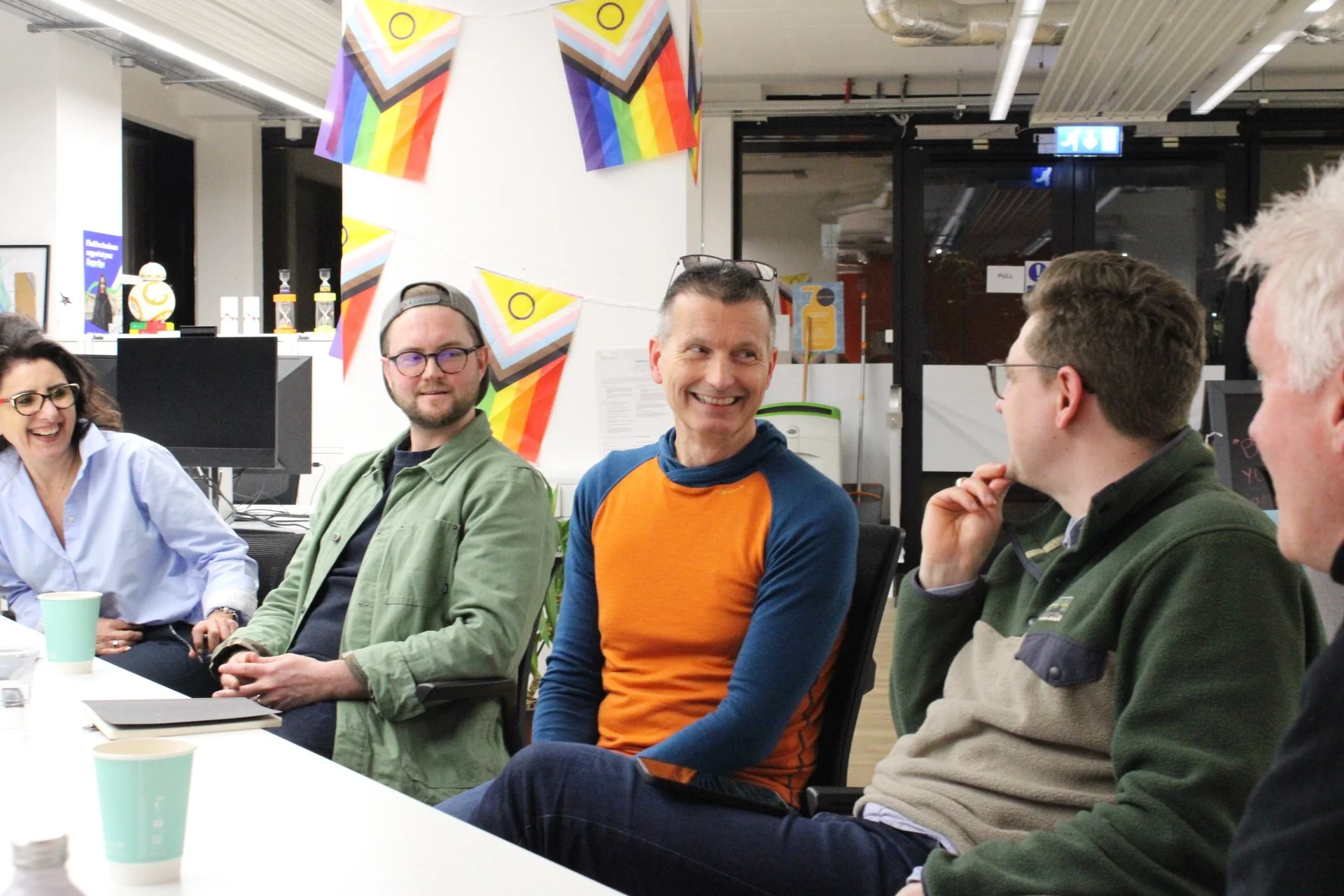 A group of five people sitting around a table at a Scottish Business Network Event, smiling and engaging in conversation, with colorful rainbow flags and decorations hanging from the ceiling.