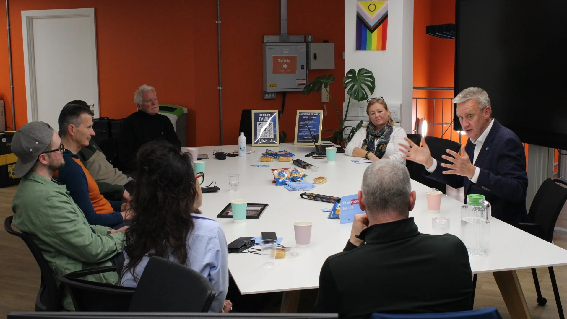 A group of people sitting around a conference table, with one man standing and gesturing as he speaks during a meeting in a modern room with orange walls, a large television screen, and decorative artwork.