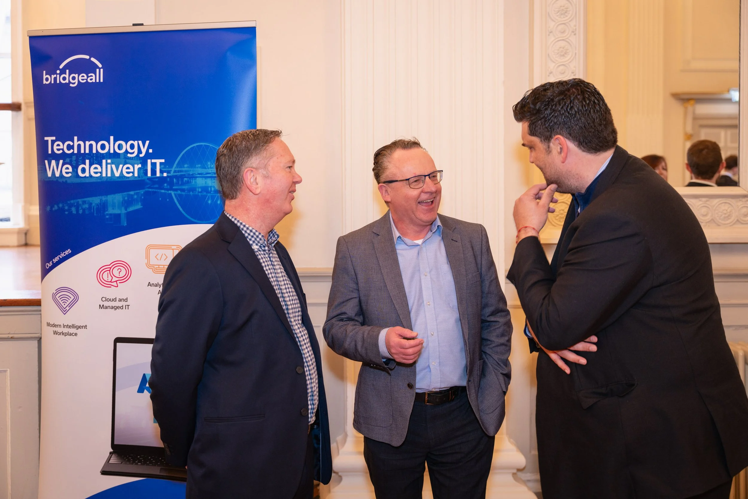 Three men in business attire are having a conversation in front of a blue banner that says 'bridgeall' and 'Technology. We deliver IT.' The setting appears to be an indoor event or conference with ornate decor and a mirror on the wall.