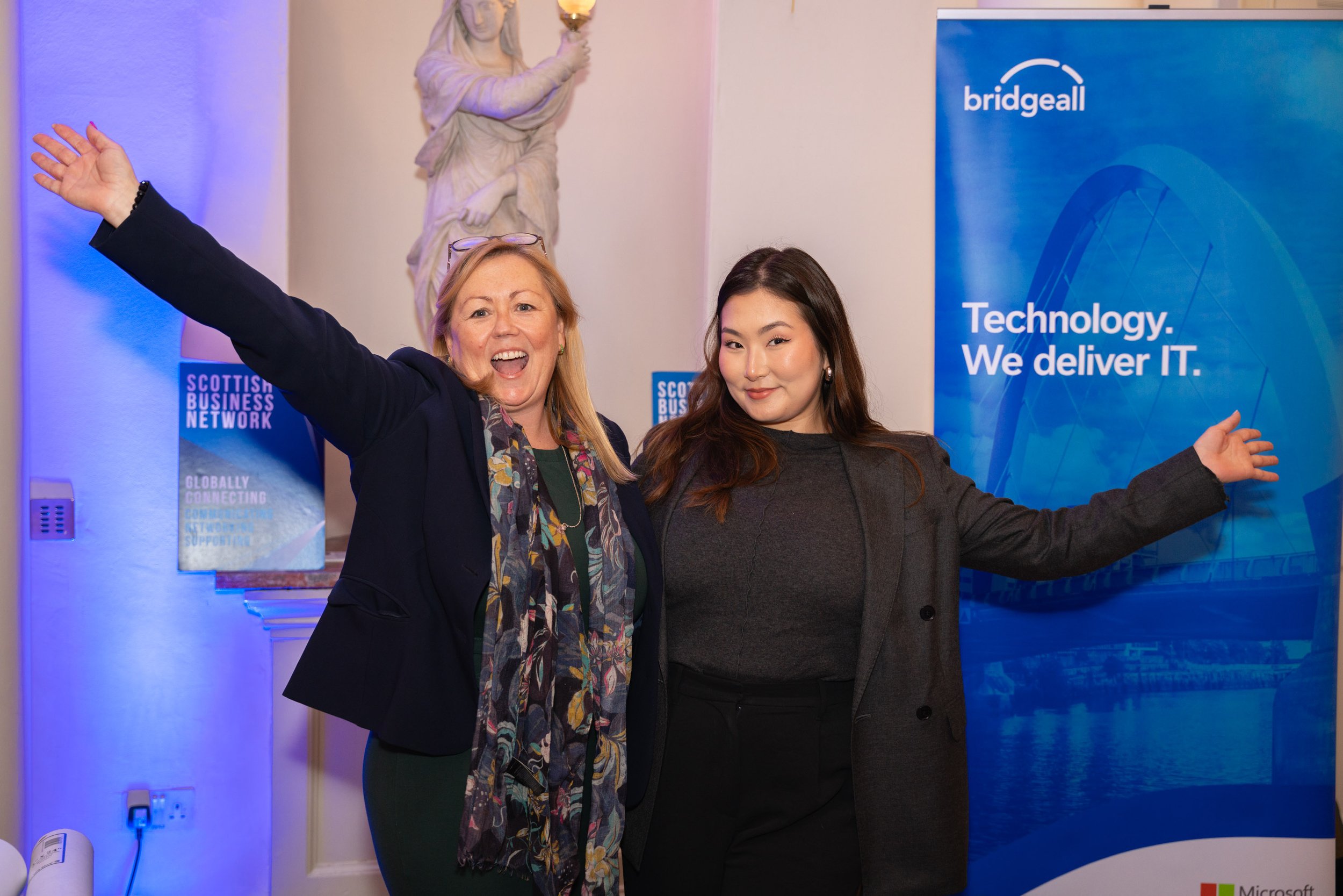 Two women standing in front of blue banners at a business event, one woman with her arms outstretched and smiling, the other with arms open, in front of a statue of a woman holding a lamp, the banners display the text 'Scottish Business Network' and 'bridgeall Technology. We deliver IT.'.