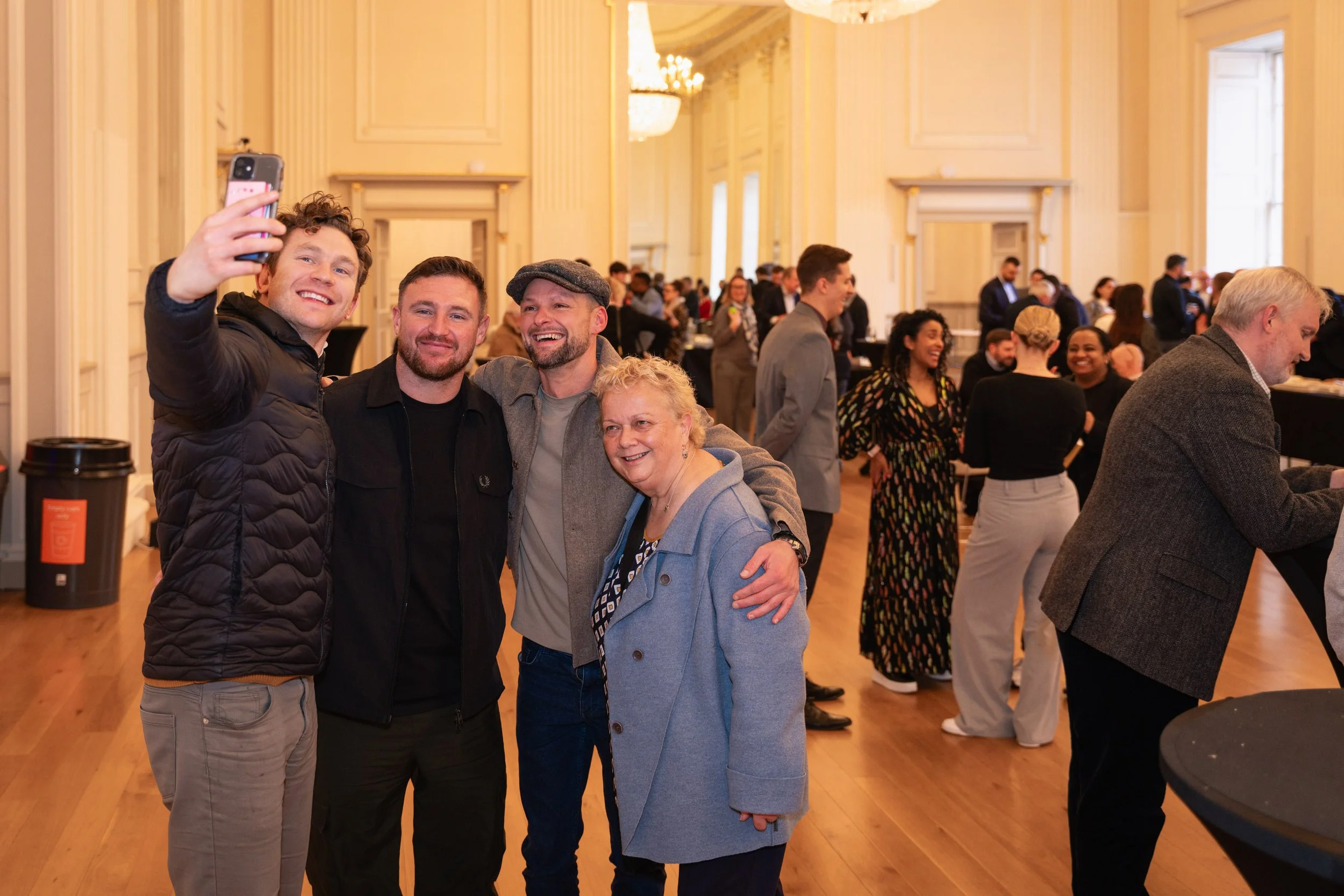 Four people taking a selfie at a social gathering in a large, elegant room with high ceilings and chandeliers, with other guests in the background.