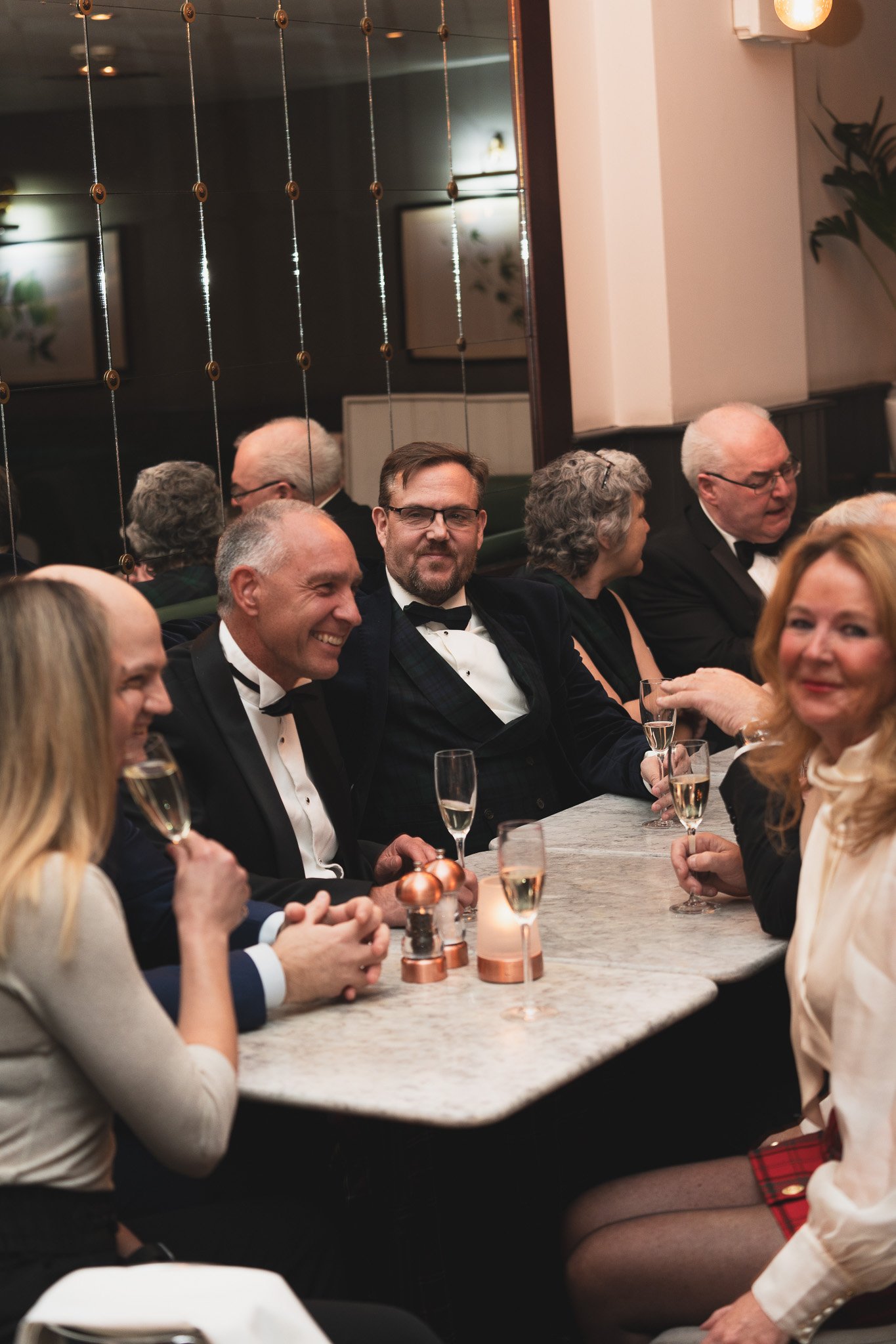A group of people dressed in formal attire, sitting at a decorated table in a restaurant or banquet hall, enjoying drinks and conversation.