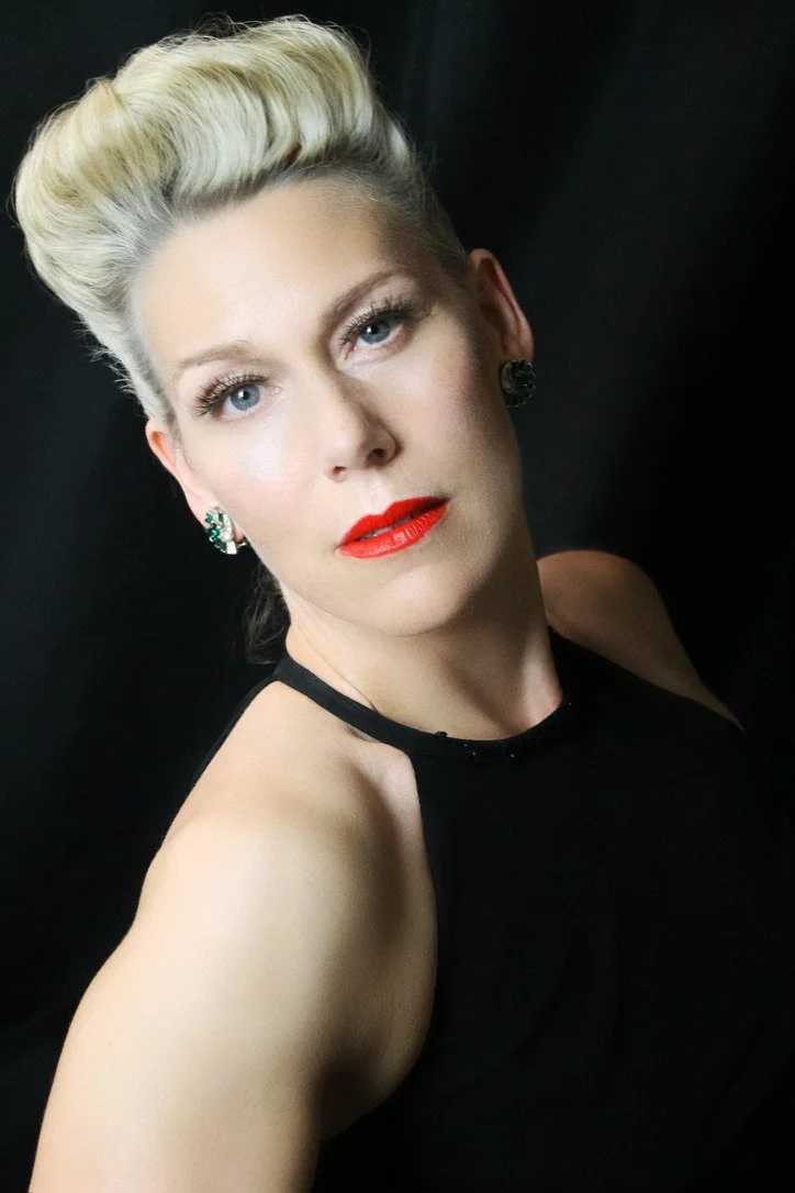 A woman with platinum blonde hair styled in an updo, wearing red lipstick, earrings, and a black sleeveless top, poses against a dark background.