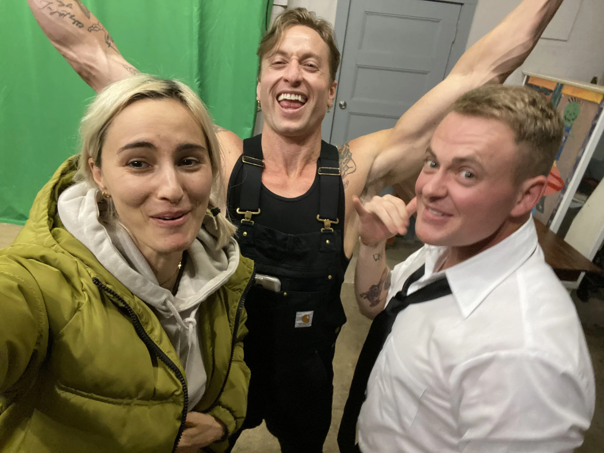 Three friends taking a selfie together indoors, with a green screen behind them. The woman on the left has blonde hair, is wearing a yellow jacket and a hoodie, and is smiling. The man in the middle has light brown hair, is wearing black overalls, an