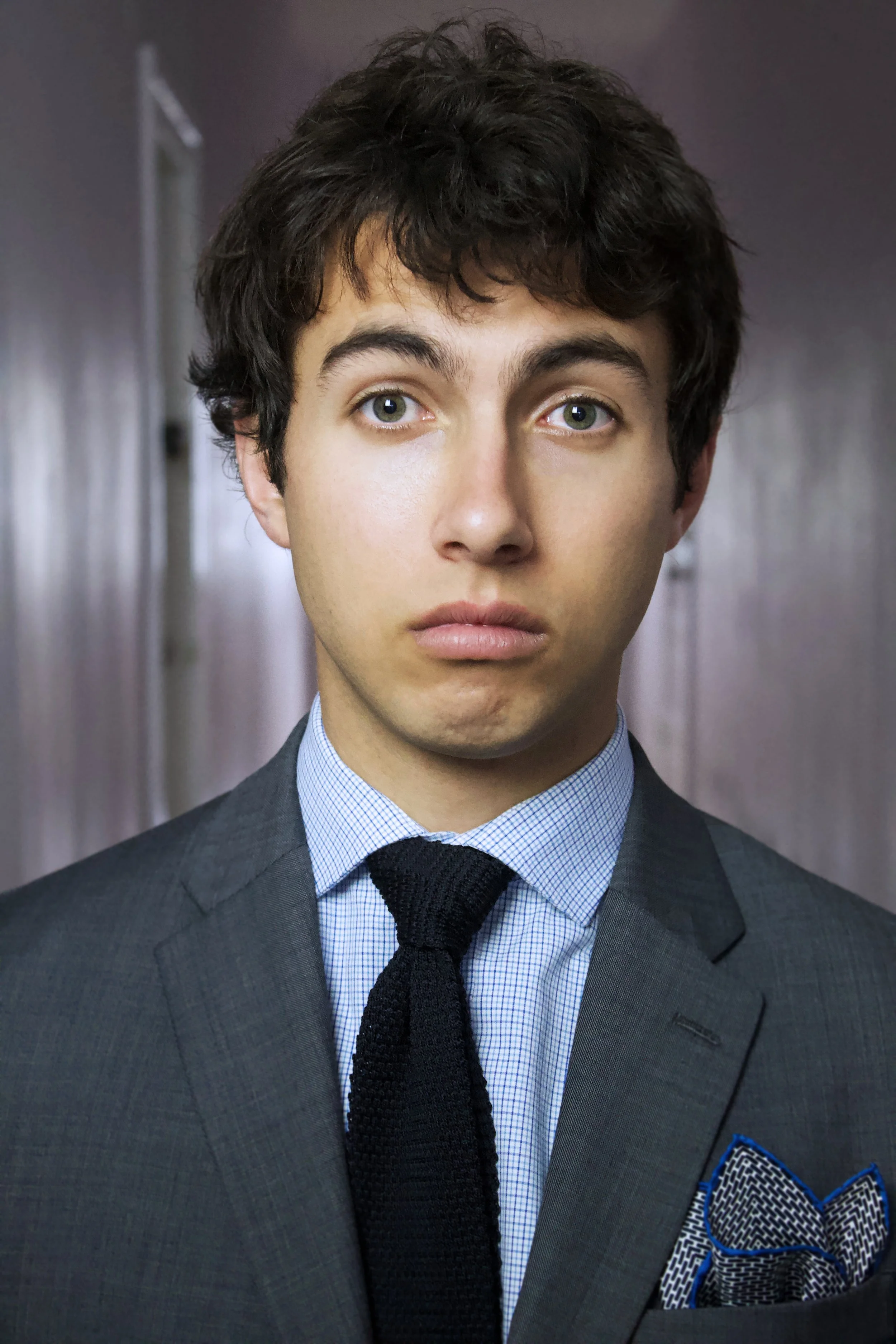 Close-up of a young man with curly brown hair, dressed in a grey suit, light blue checked shirt, and black tie, standing in a hallway with a blurred background.