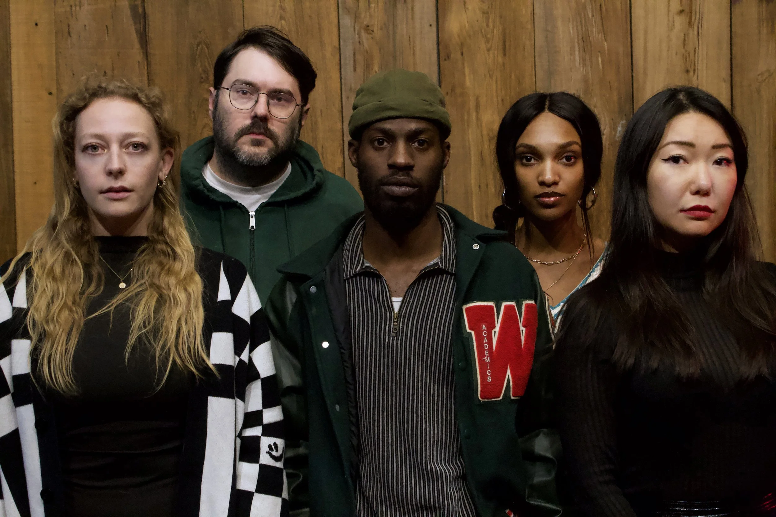 Five young adults standing together in front of a wooden wall, looking serious.